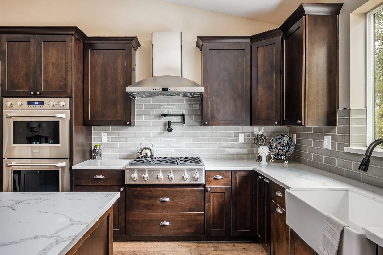 Professional gas cooktop with pot filler, chimney hood, and double wall ovens in dark wood cabinetry
