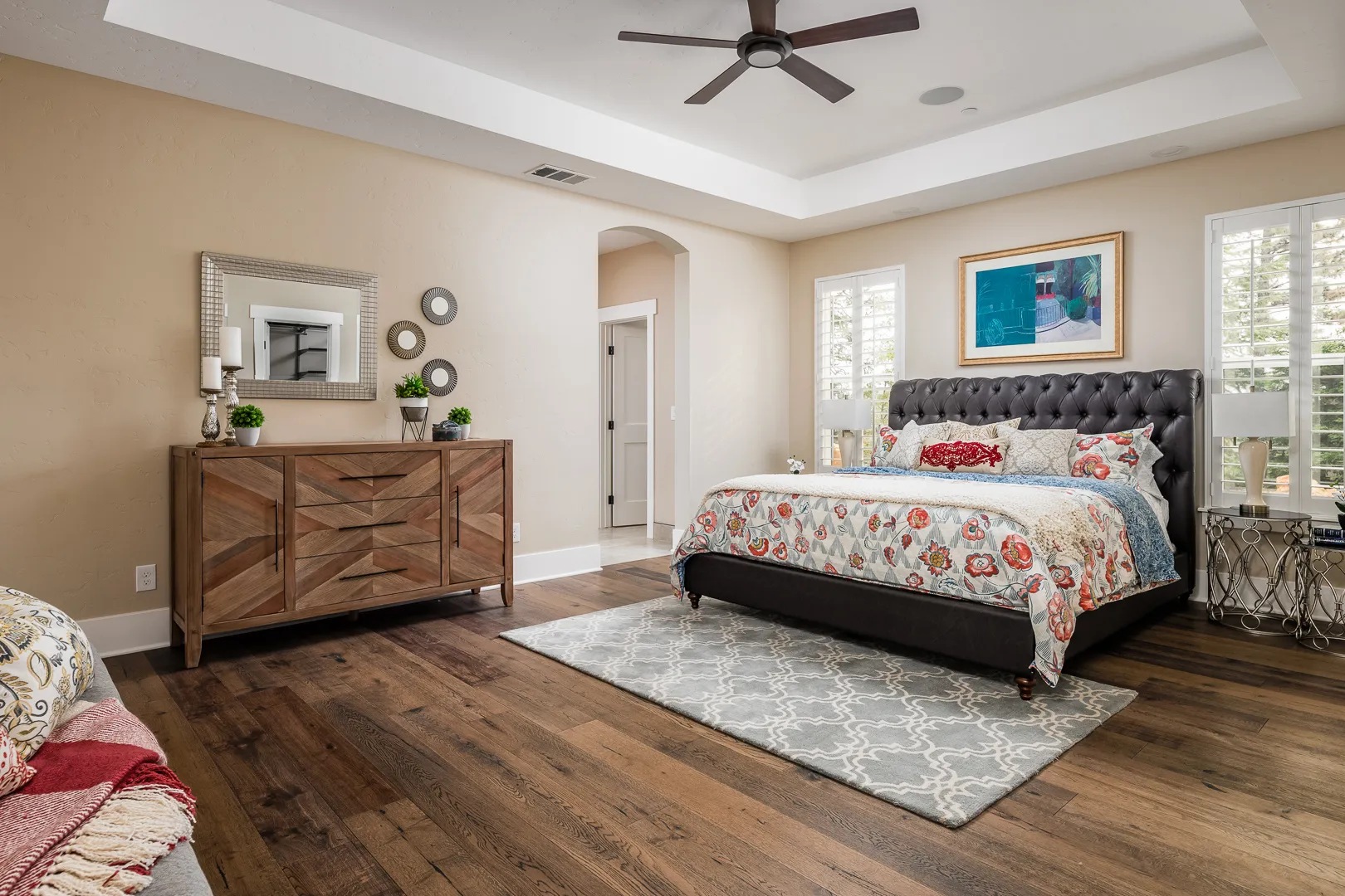 Primary bedroom with tray ceiling, ceiling fan, tufted leather bed, and hardwood floors