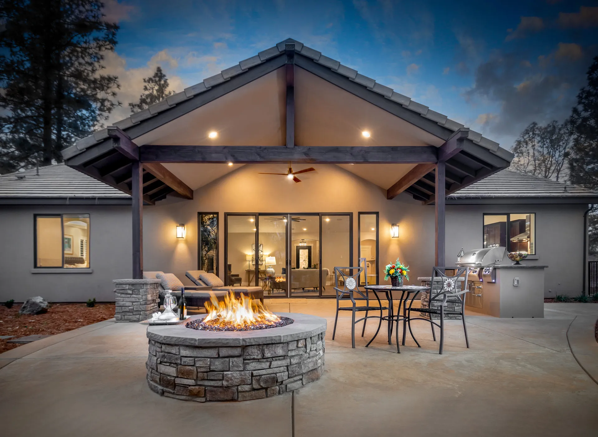 Covered timber-frame patio at dusk with stone fire pit, bistro seating, and built-in grill