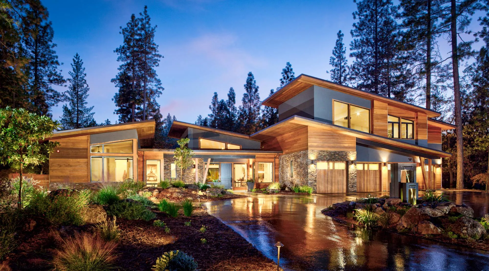 Modern cedar, stone and glass home at twilight among the pines, with lit windows and a wet driveway reflecting the light