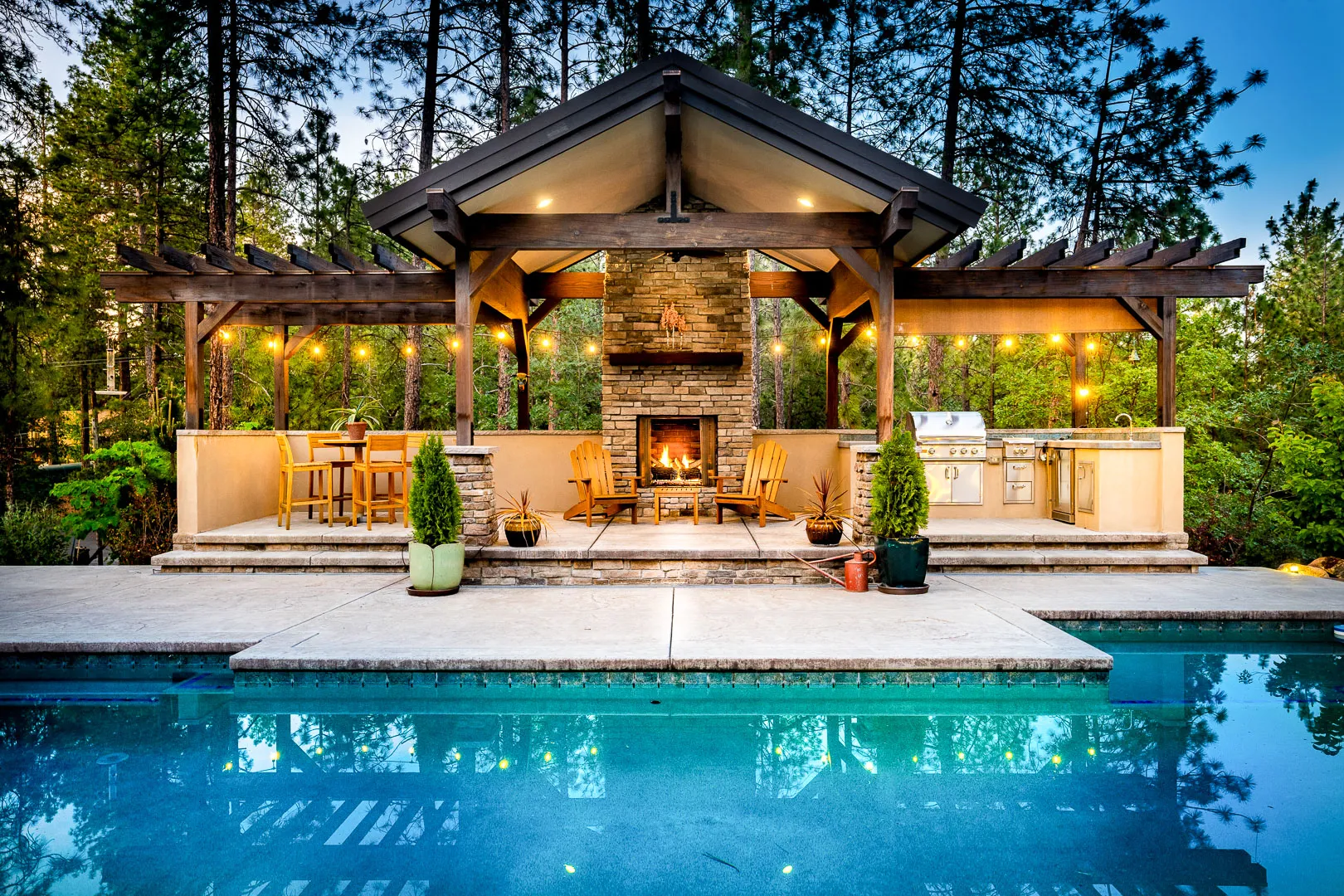 Post-and-beam outdoor pavilion with stone fireplace and BBQ island beside a pool