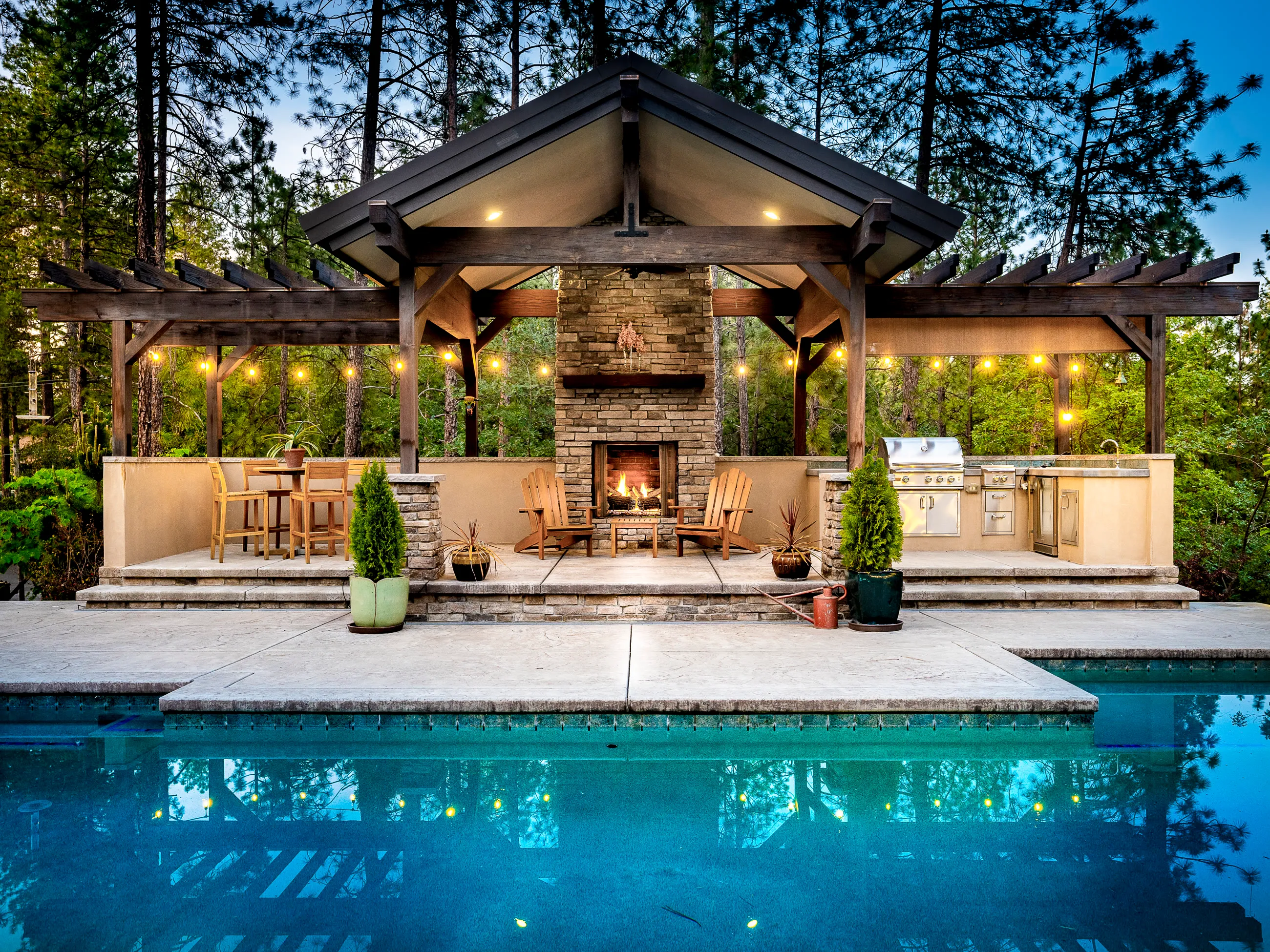 The same pavilion finished — stone fireplace, string lights, and the pool glowing at dusk