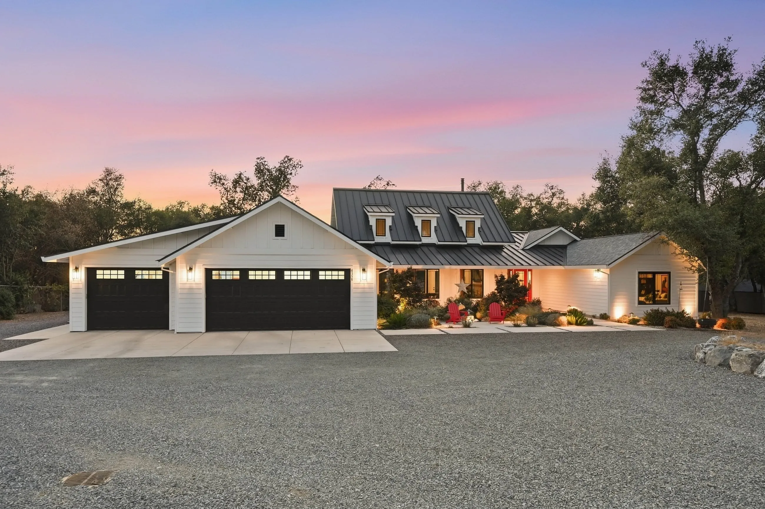 Front elevation at dusk with three-car garage wing, dormers, and lit landscaping