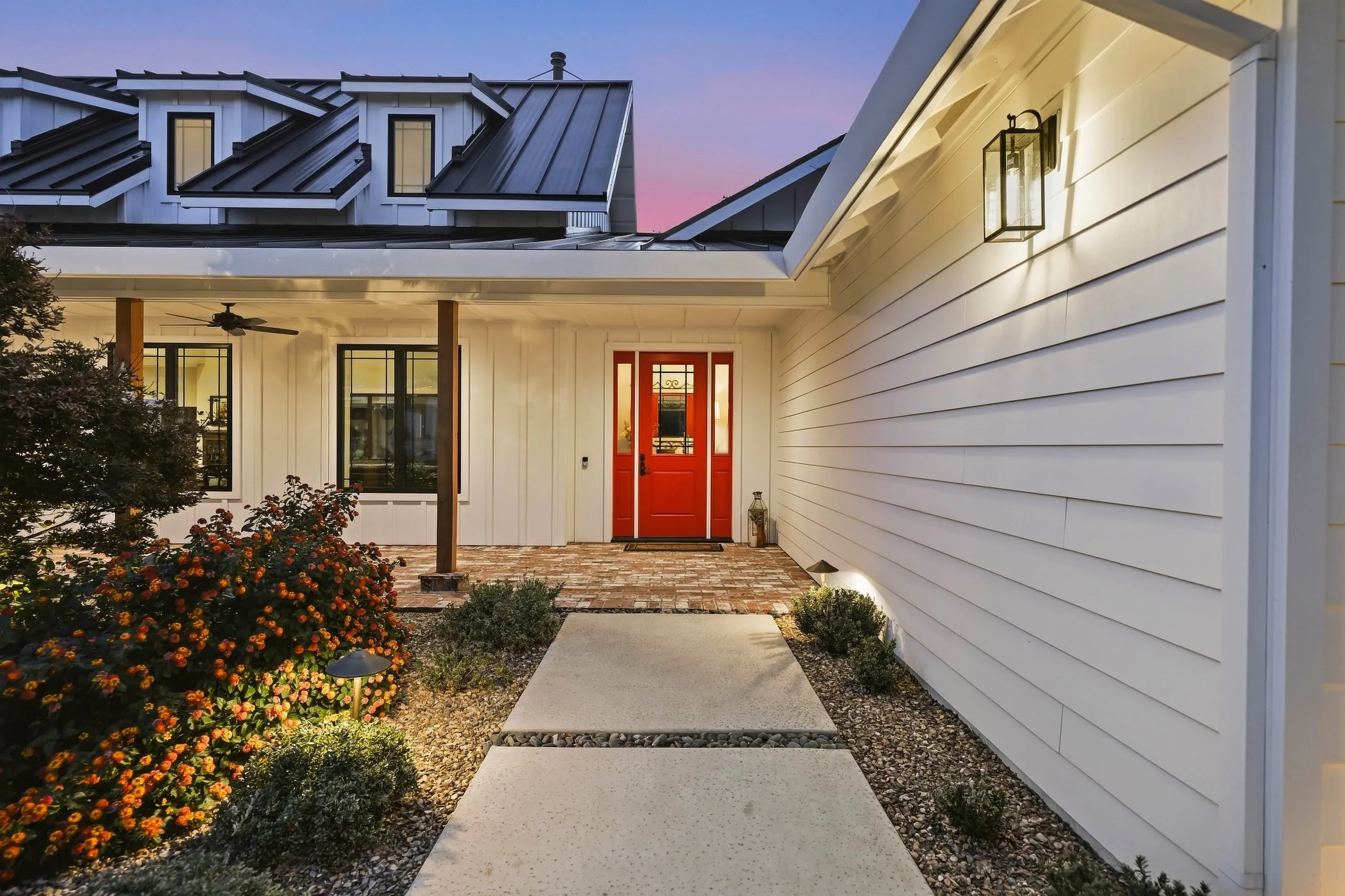 Entry walk to the red front door with lantern sconces and standing-seam dormers at dusk