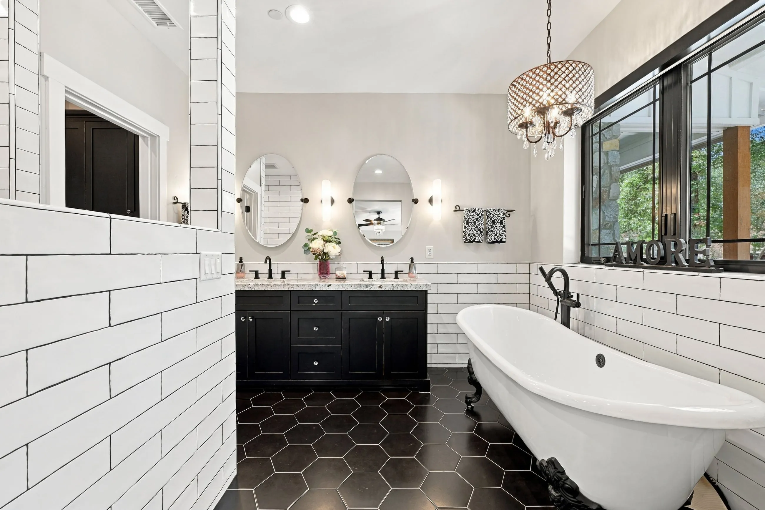 Primary bath with black clawfoot tub, double vanity, oval mirrors, and beaded chandelier