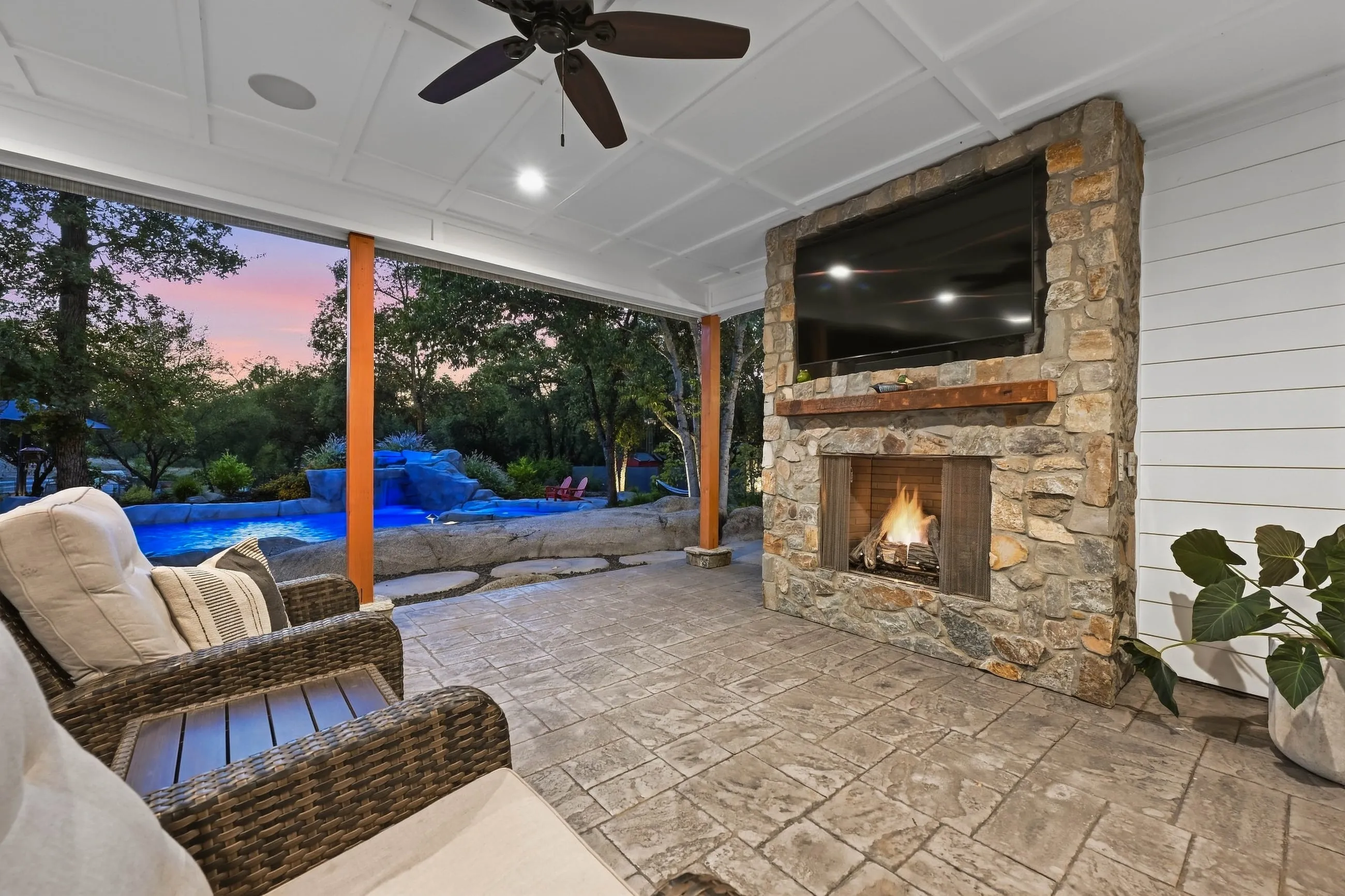 Outdoor stone fireplace under the porch with the lit pool beyond at dusk
