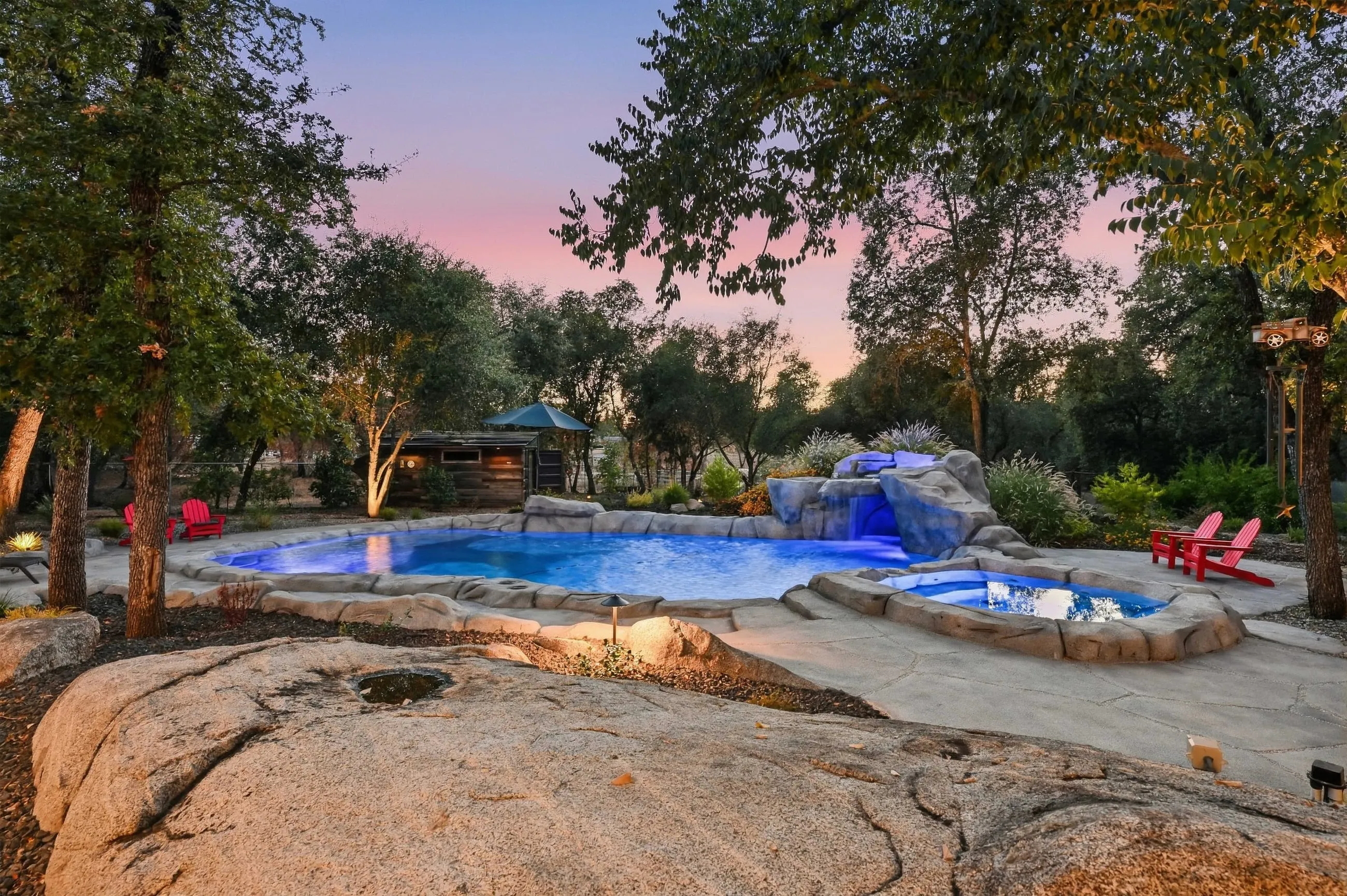 Pool and spa lit blue at twilight with rock waterfall and red chairs