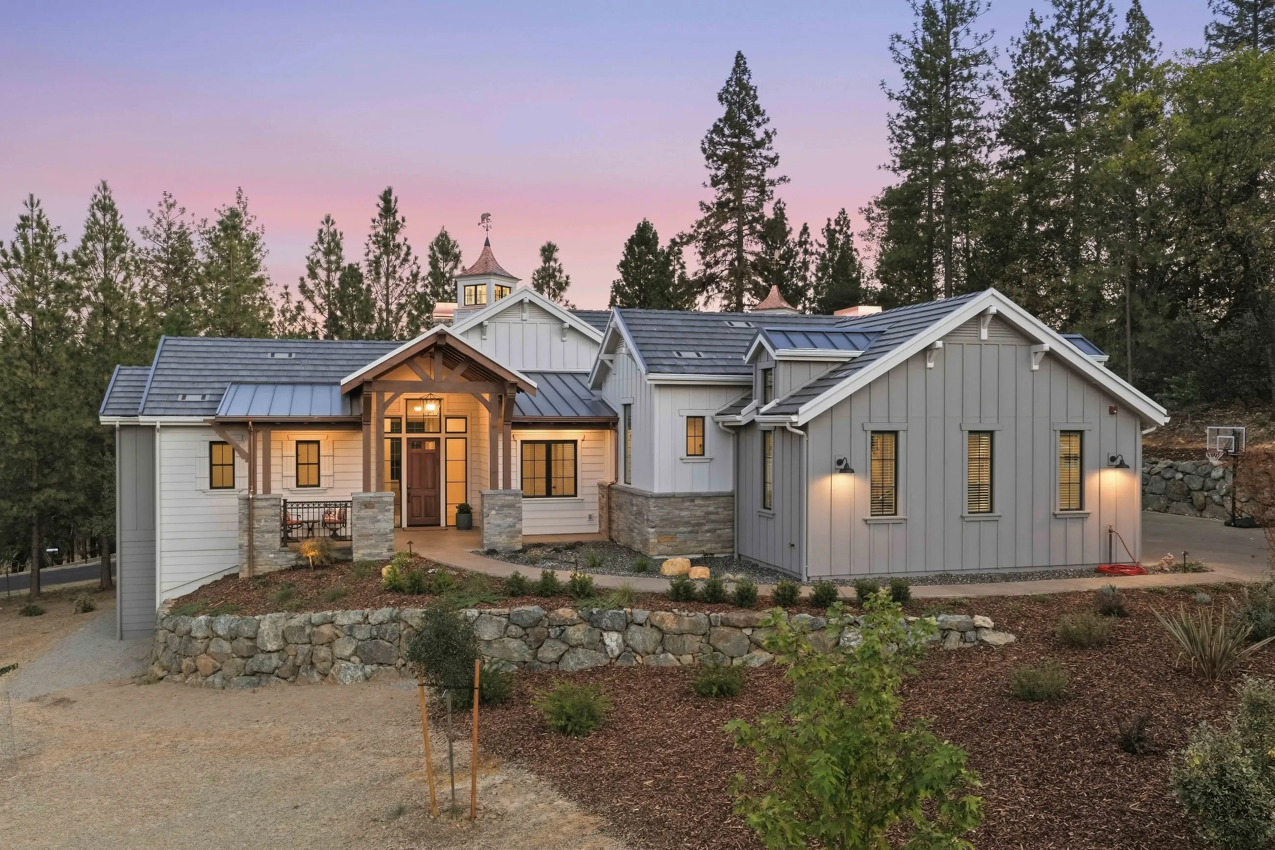 Front elevation at twilight with timber entry portico, stone retaining walls, and warmly lit windows