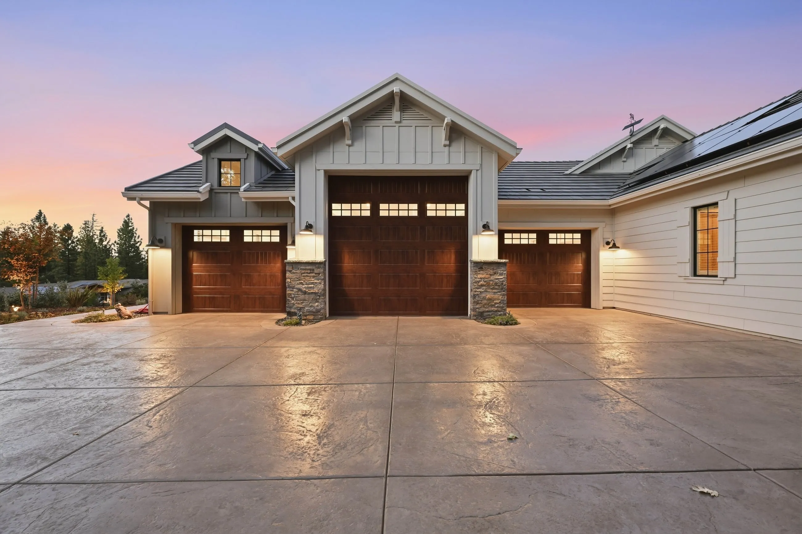 Garage wing with three carriage-style wood doors and stone piers glowing at dusk