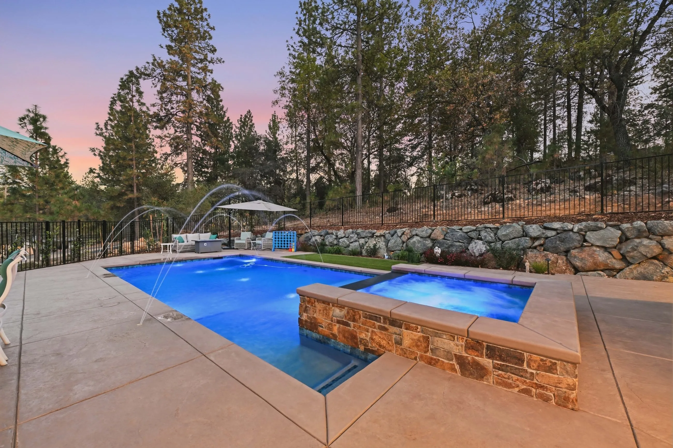 Raised stone spa spilling into the pool at twilight beneath a boulder retaining wall