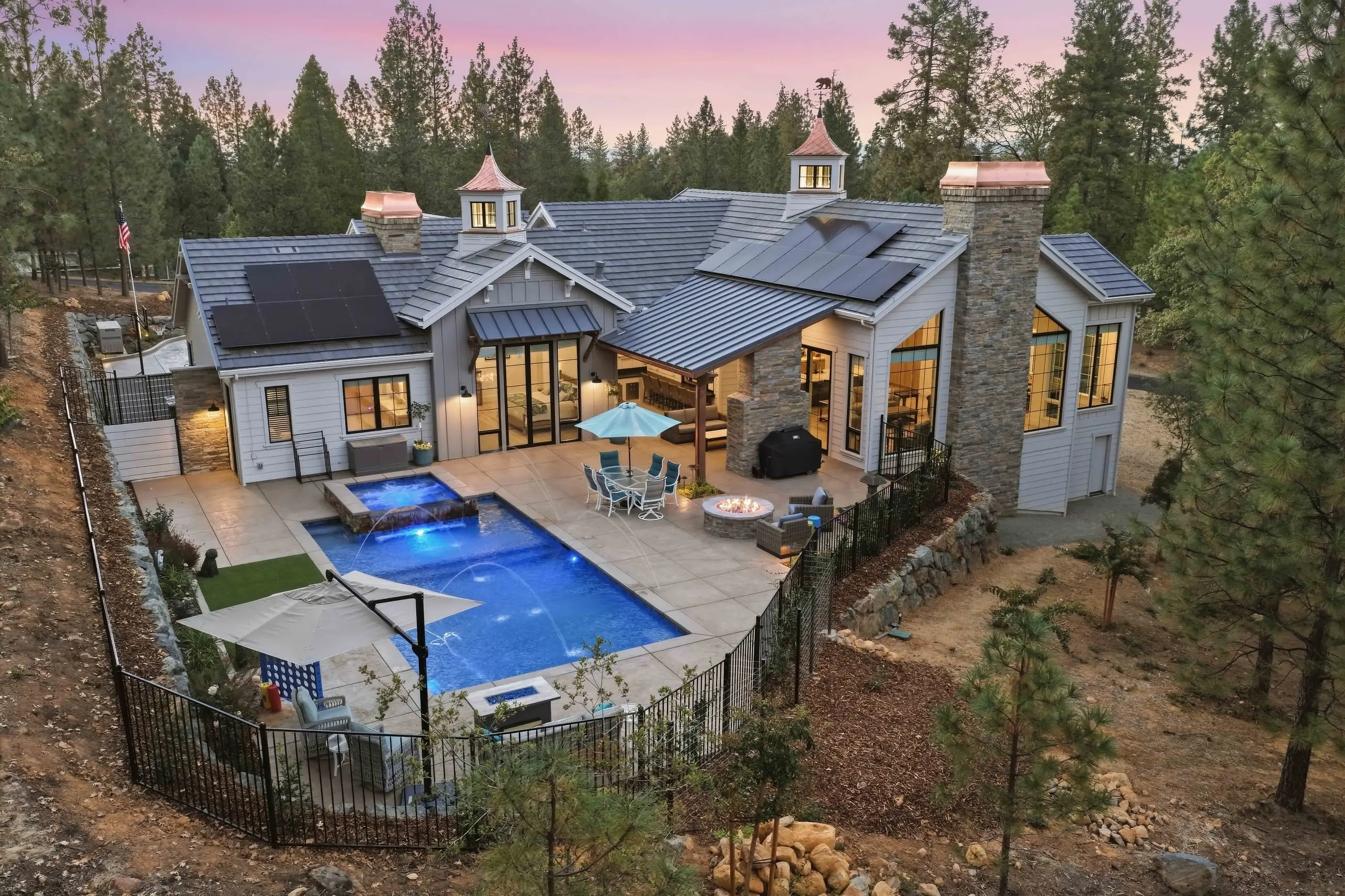 Aerial twilight view of a Sherman modern farmhouse backyard — pool with water arcs, spa, fire pit, and outdoor living among the pines