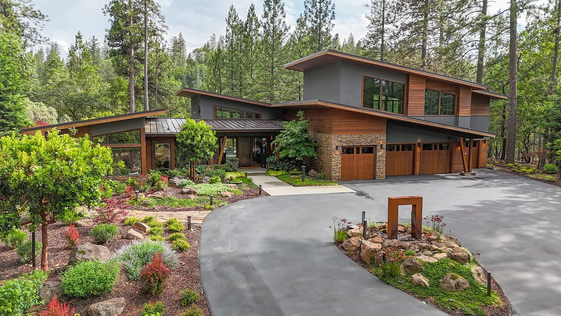 Front of the mountain-modern home with cedar siding, stacked stone, three wood garage doors and a curving drive through drought-tolerant gardens
