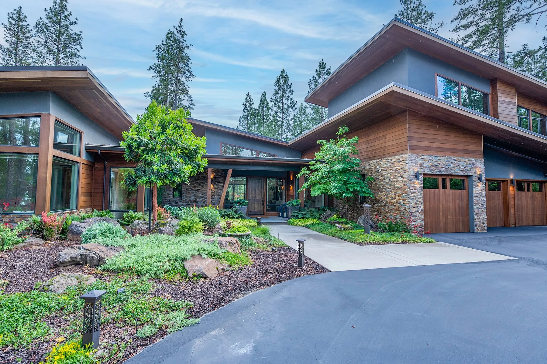 Covered entry walk framed by maples and layered plantings, with the two-story cedar and stone wing beyond