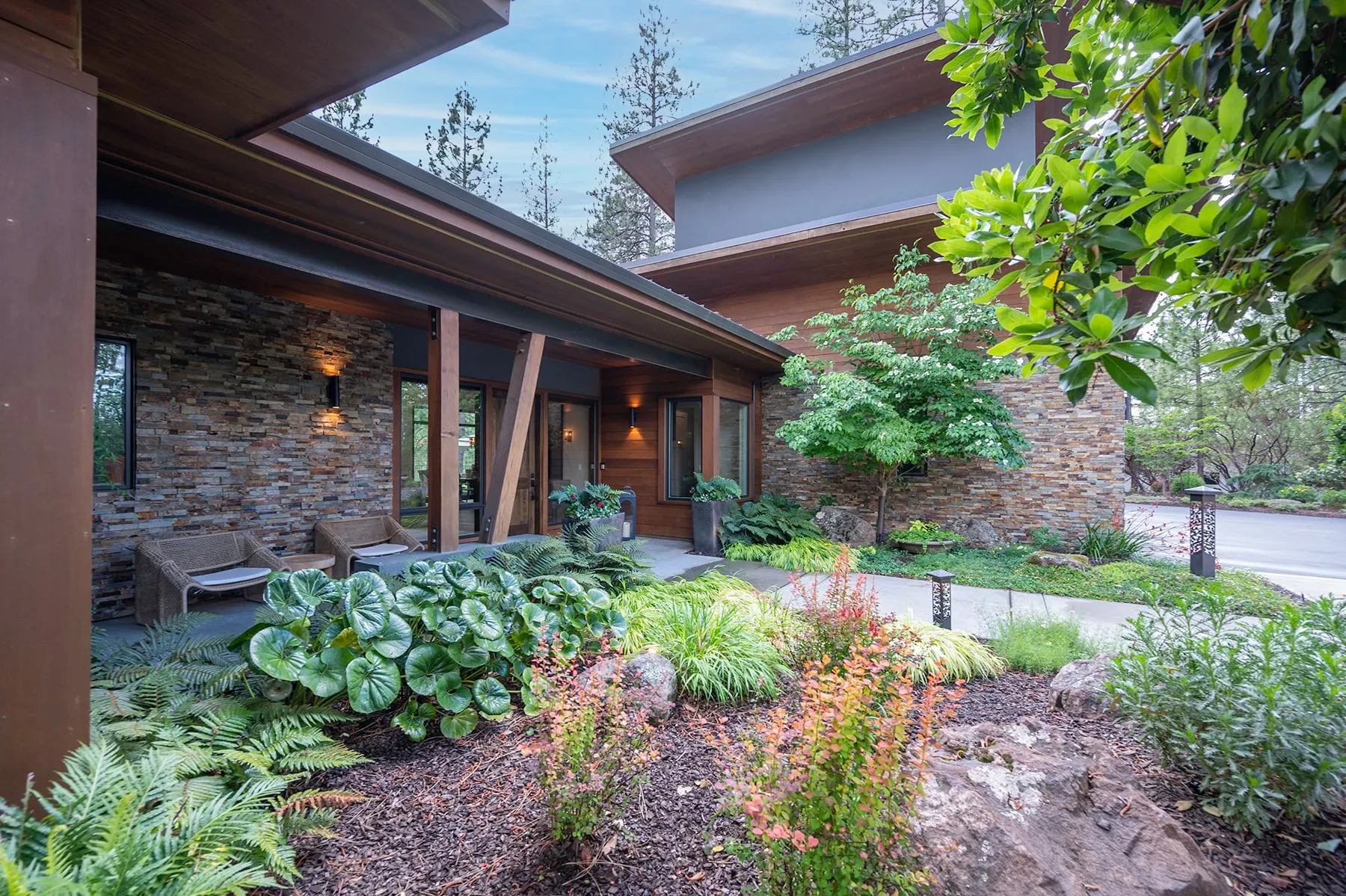 Angled view of the covered entry porch with cedar soffits, stone walls and lush shade plantings