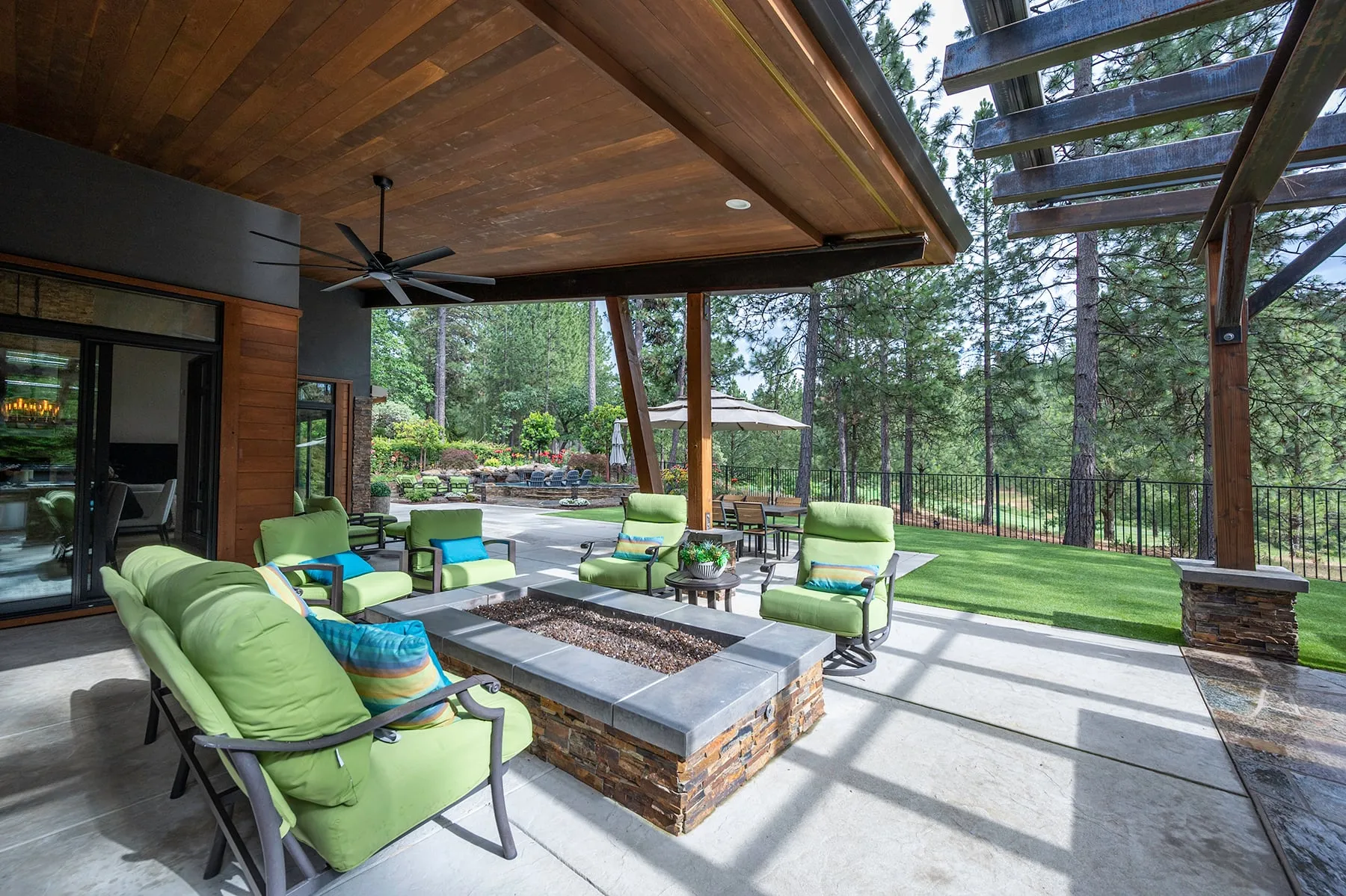 Covered porch around a stacked-stone fire pit table, opening to a pergola and the fenced lawn