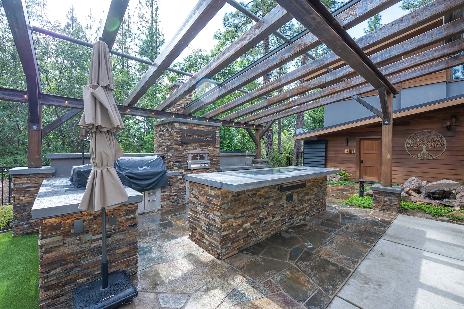 Outdoor kitchen under a steel and timber pergola with stone-clad counters, a grill and a pizza oven