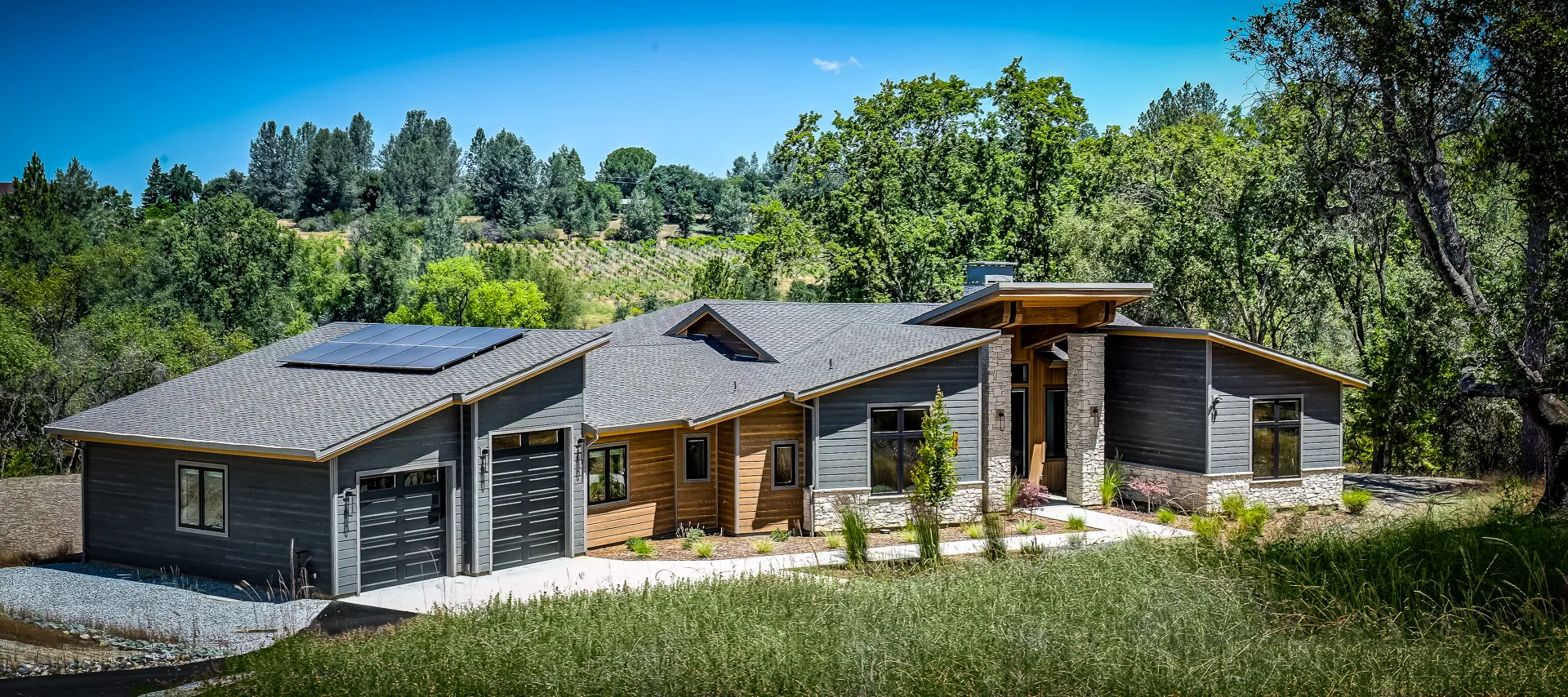 Front exterior with charcoal and cedar-tone siding, solar panels, and a three-car garage under blue skies