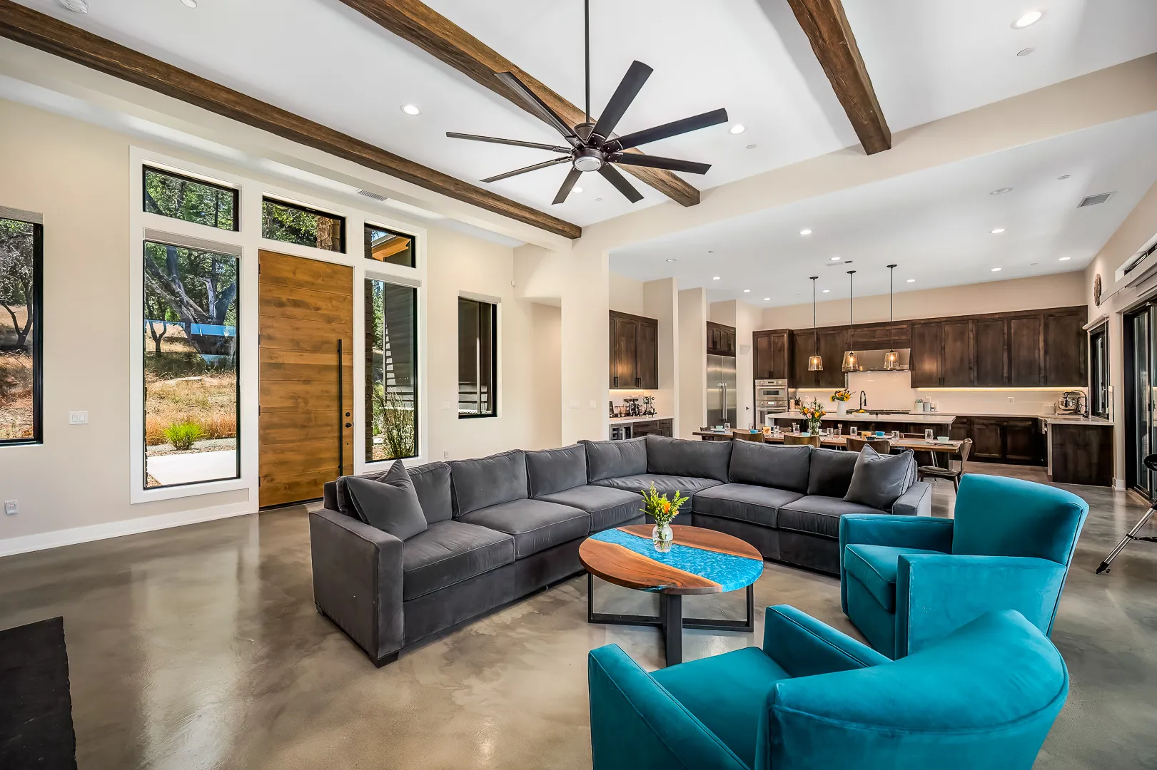 Great room with rustic ceiling beams, gray sectional, teal armchairs, and open kitchen beyond