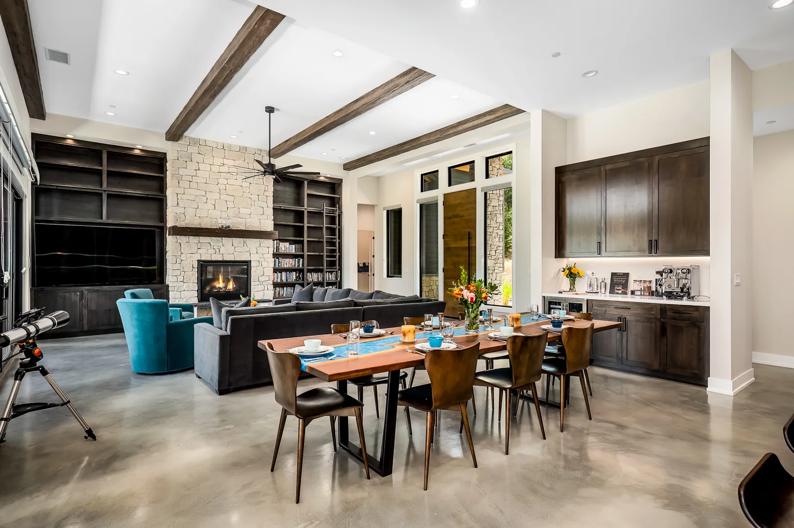 Dining area with live-edge table, coffee bar cabinetry, stone fireplace, and beamed ceiling