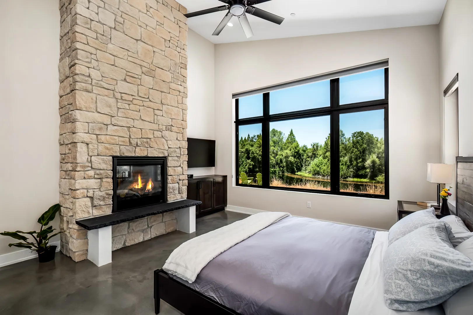 Primary bedroom with stone fireplace, ceiling fan, and a wide picture window on the pond