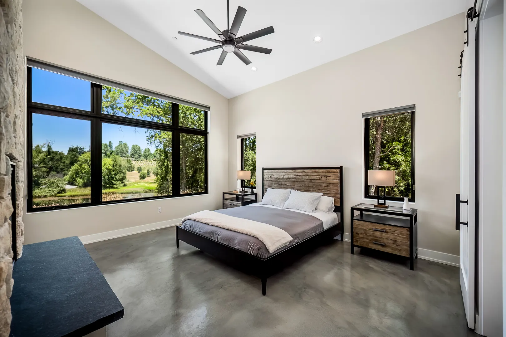 Bedroom with big black-framed corner windows overlooking trees and meadow, polished concrete floor