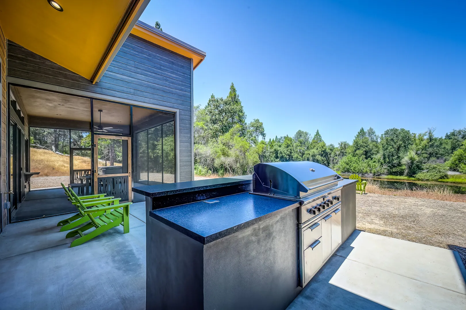 Outdoor kitchen with built-in grill and concrete counters, green Adirondack chairs on the screened porch