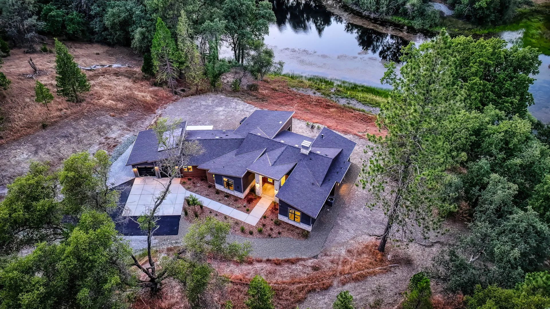Dusk aerial over the rooflines showing the home's private clearing beside the tree-lined pond