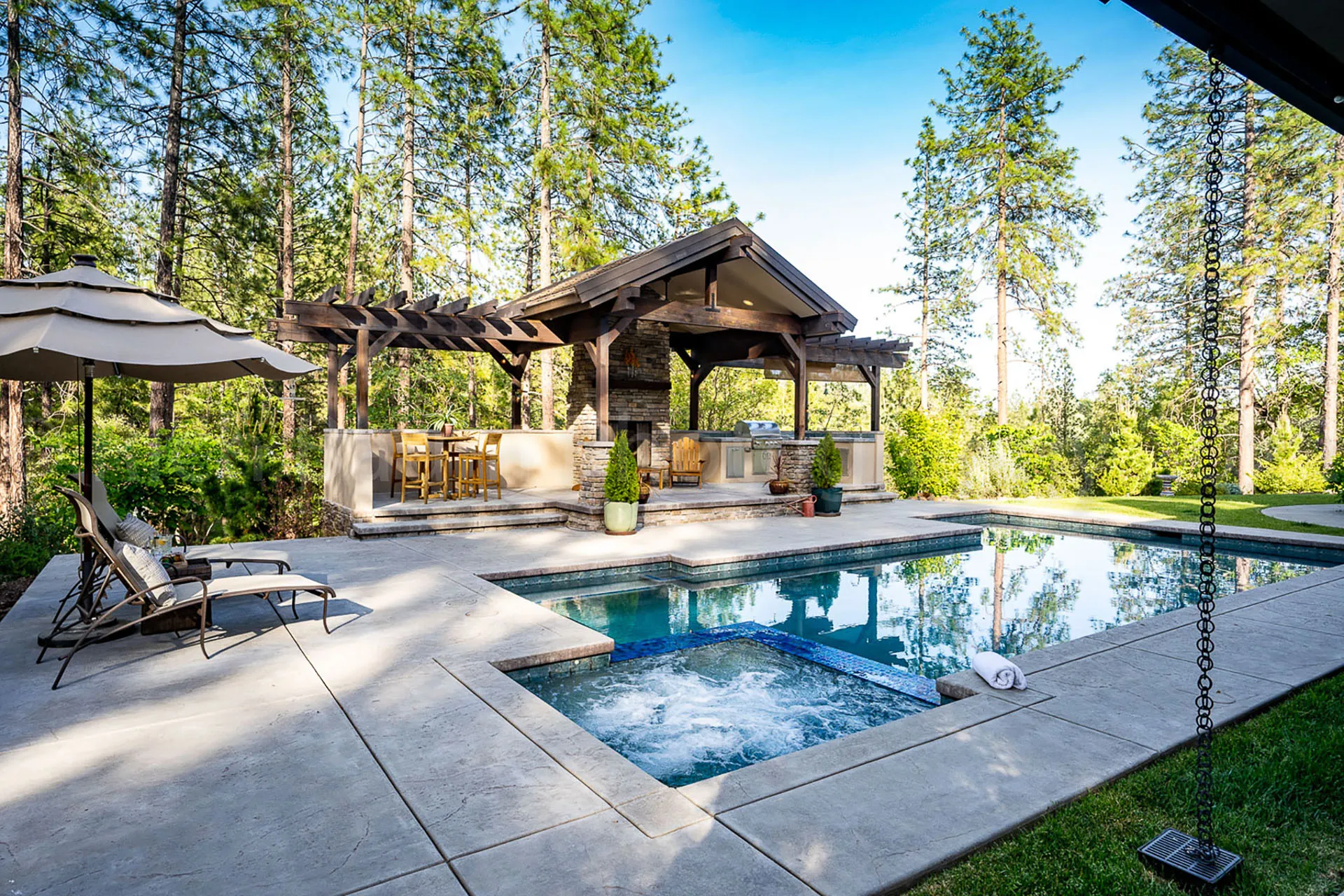 Backyard pool and bubbling spa beside a timber pavilion with outdoor kitchen among the pines