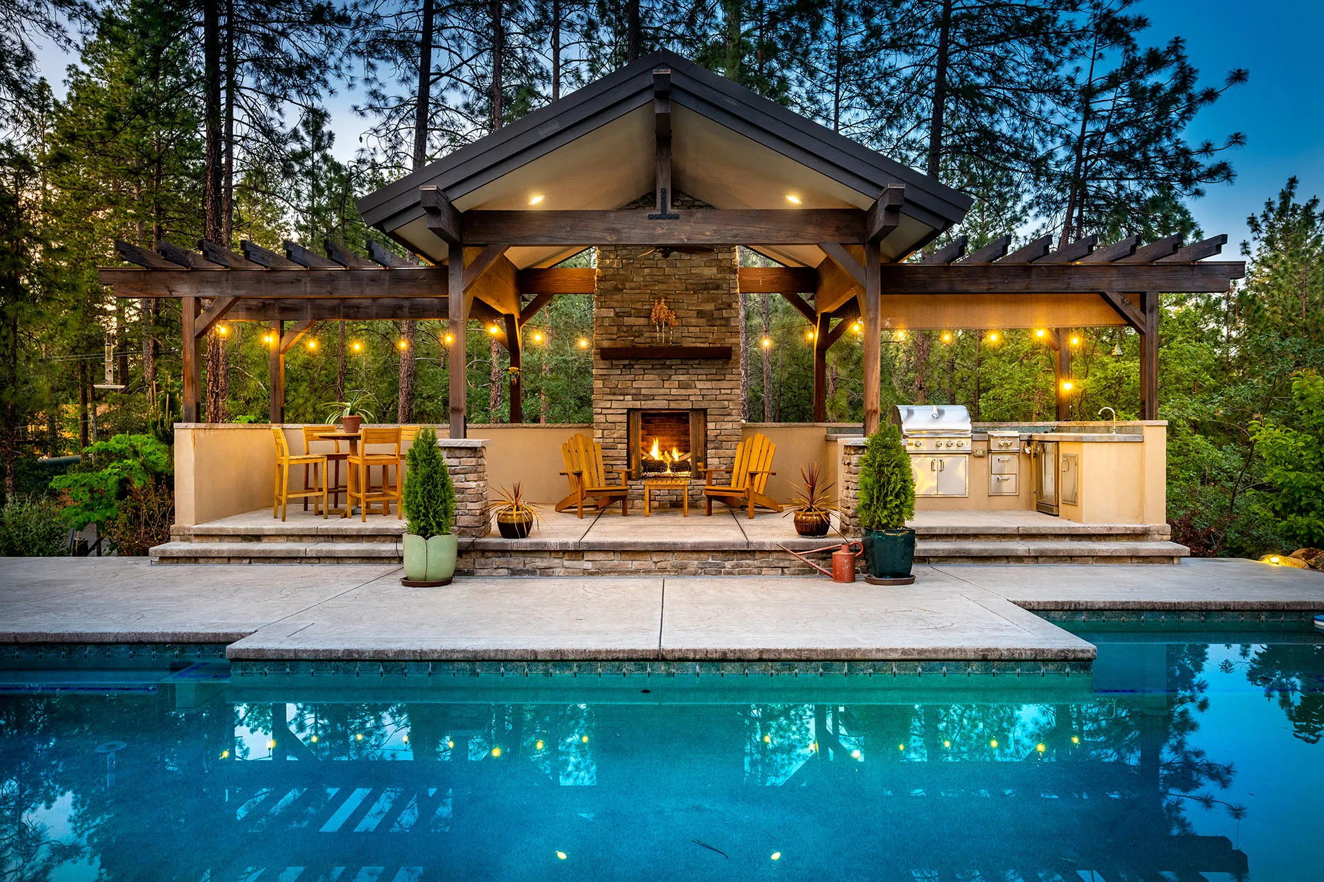 Poolside pavilion at dusk with stone outdoor fireplace, Adirondack chairs, string lights, and built-in grill