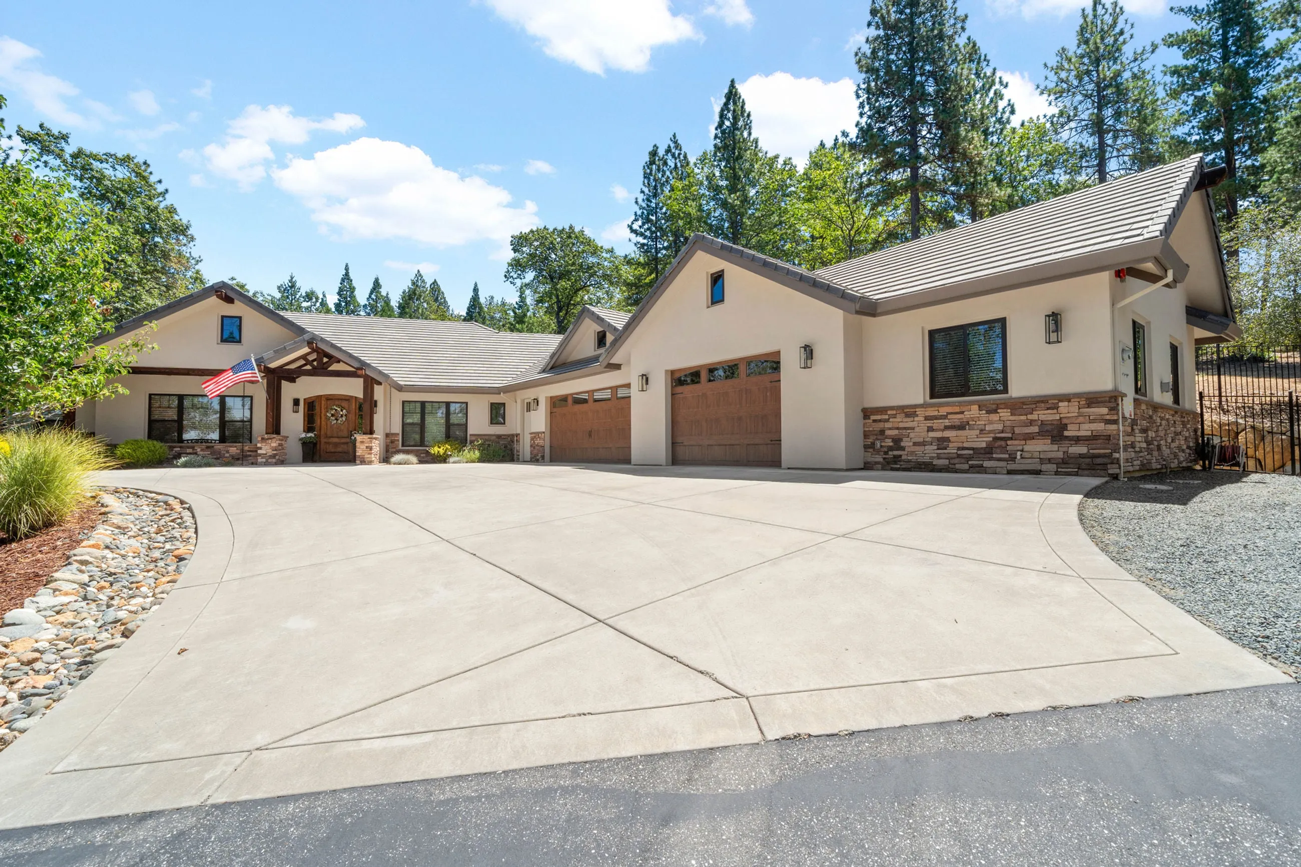 Front elevation by day with a wide concrete motor court, timber entry portico, and tall pines behind