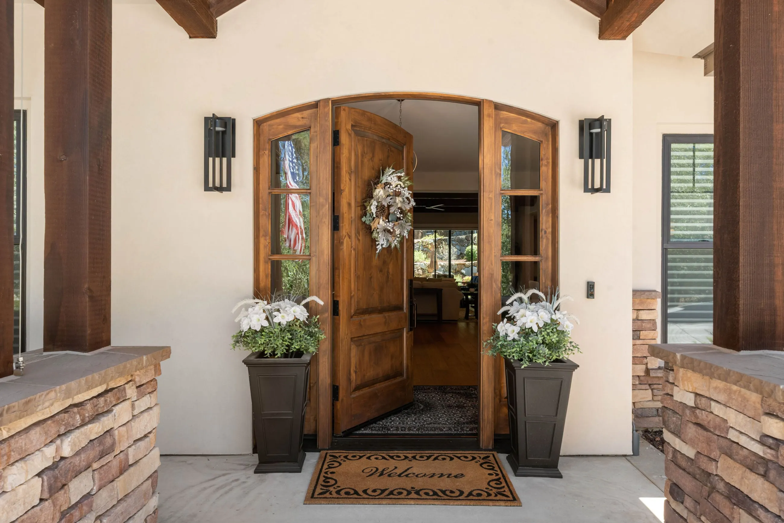 Arched alder double doors with sidelights, black lantern sconces, and planters on stone piers