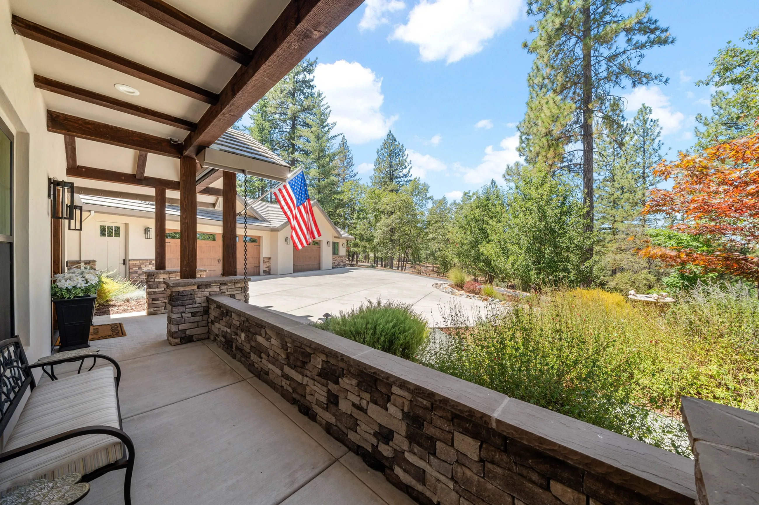 Covered front porch with a stone half wall and bench looking over the motor court and pines