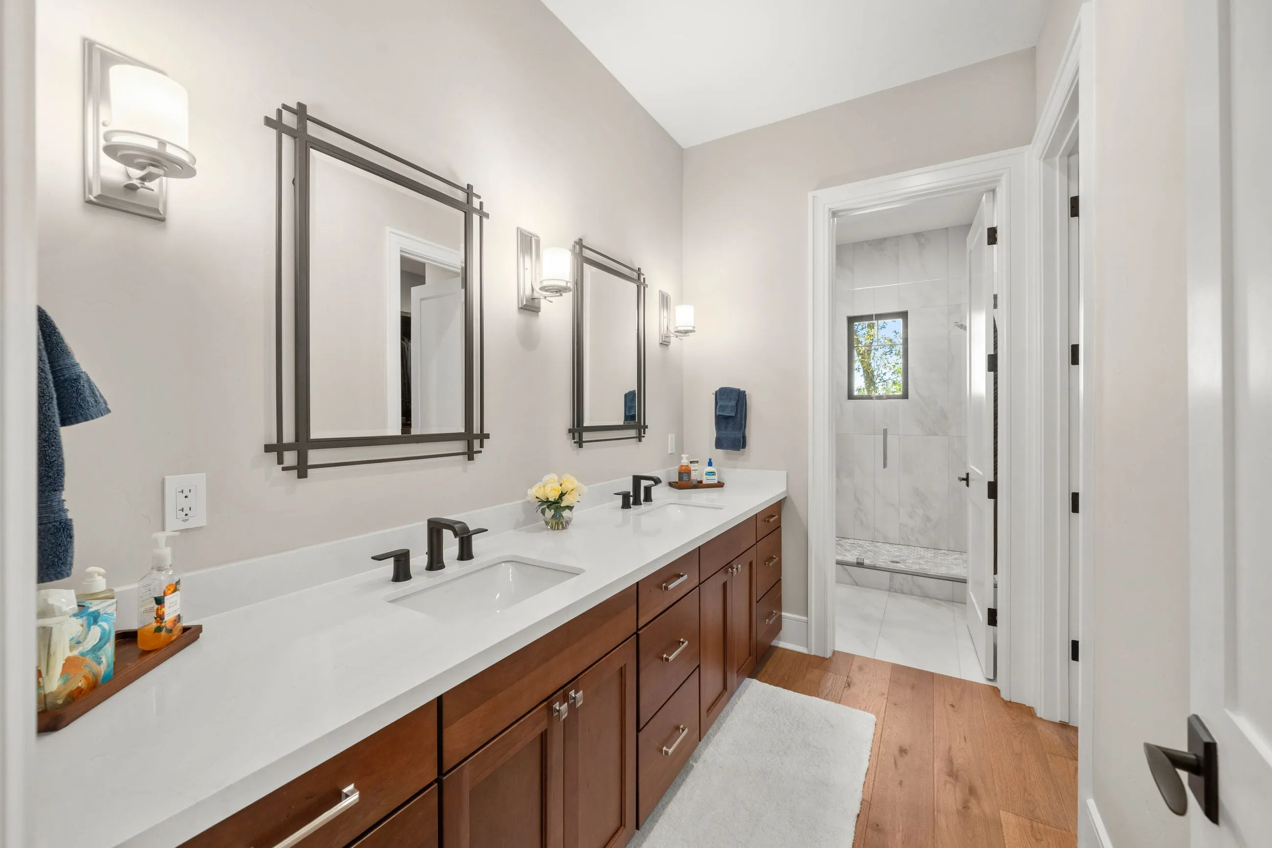 Primary bath with a long cherry double vanity, geometric mirrors, and a marble-look shower beyond
