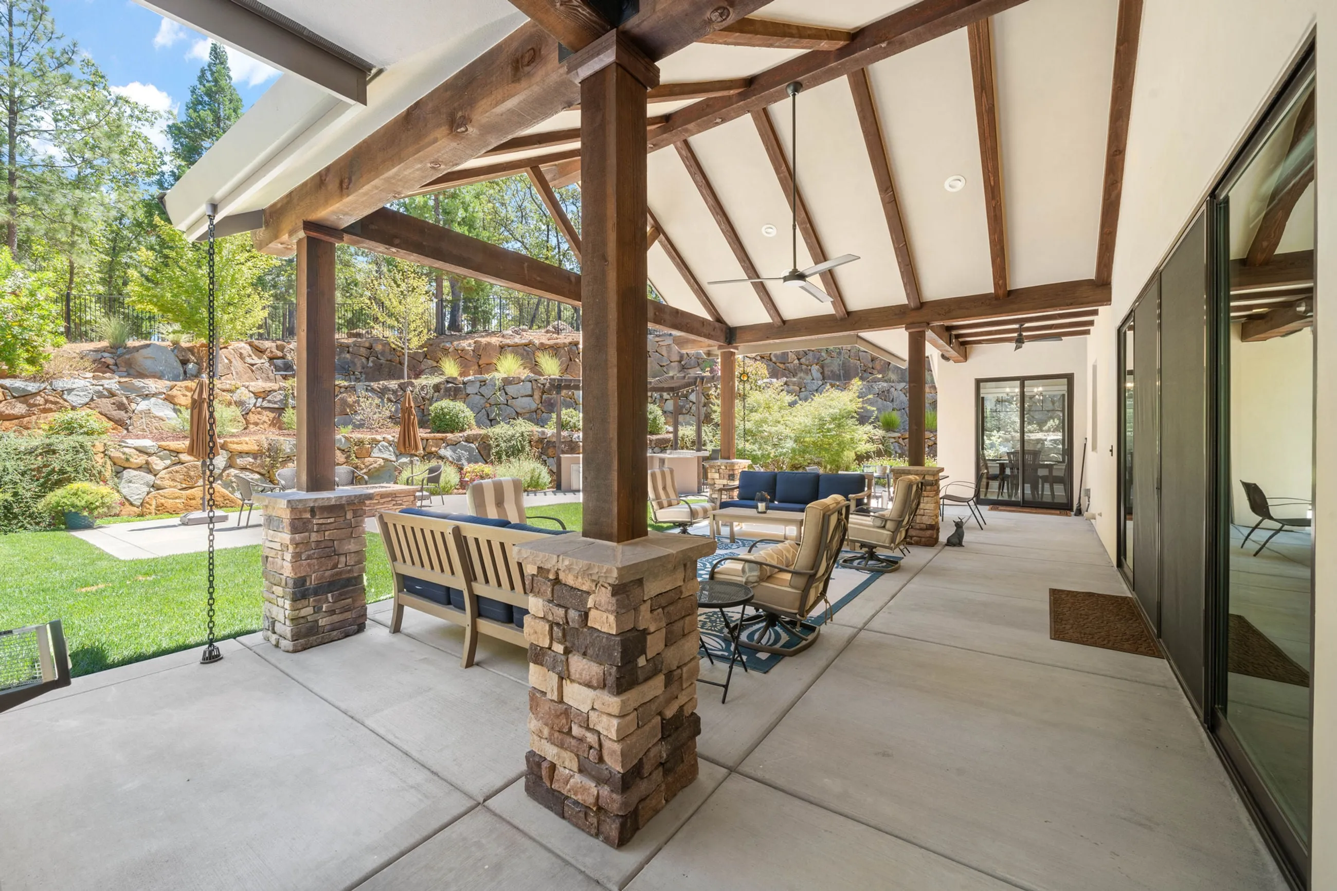 Covered patio with stacked-stone column bases, timber trusses, and seating facing the boulder wall