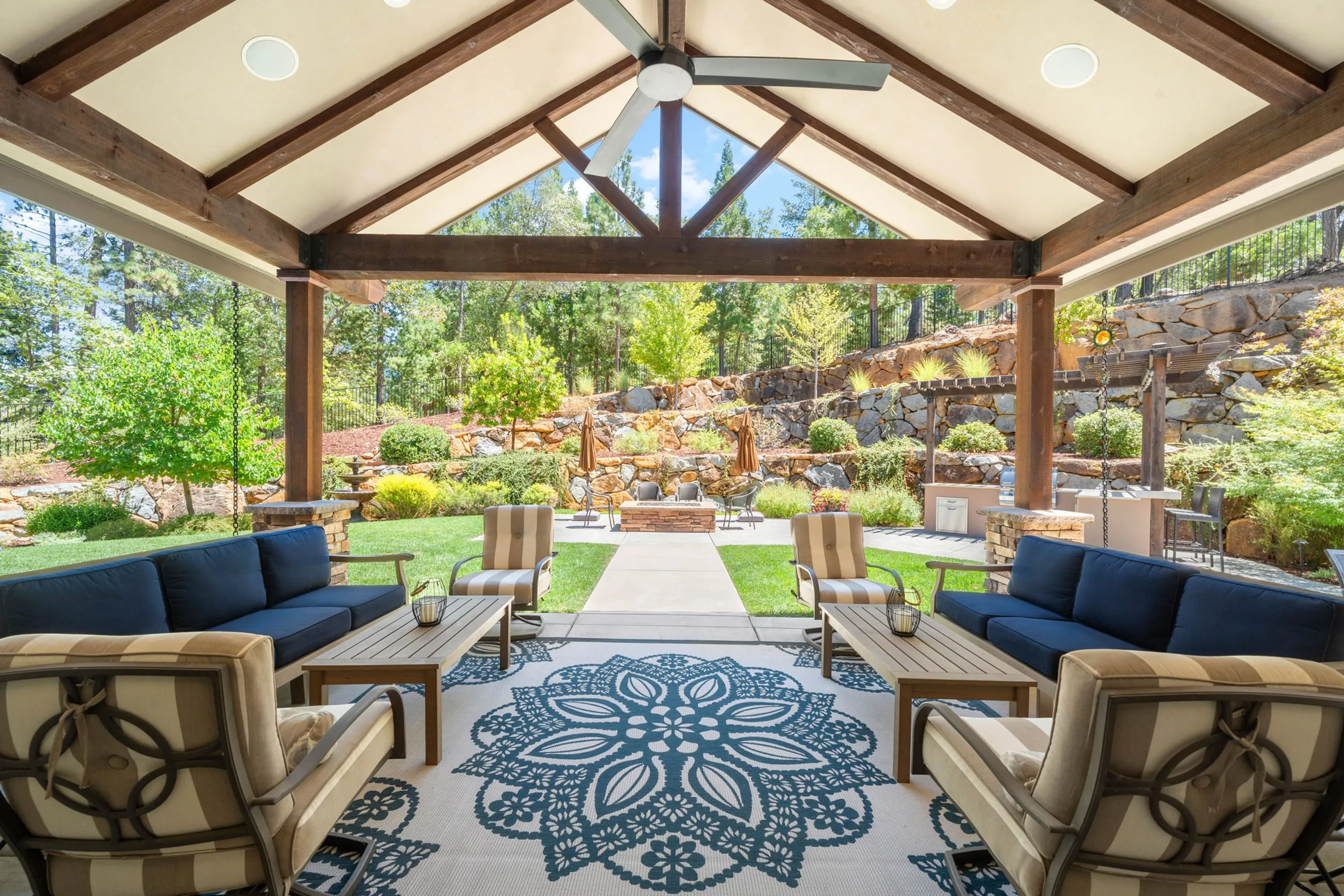 Covered outdoor living room with timber trusses, ceiling fan, and navy sofas overlooking the lawn
