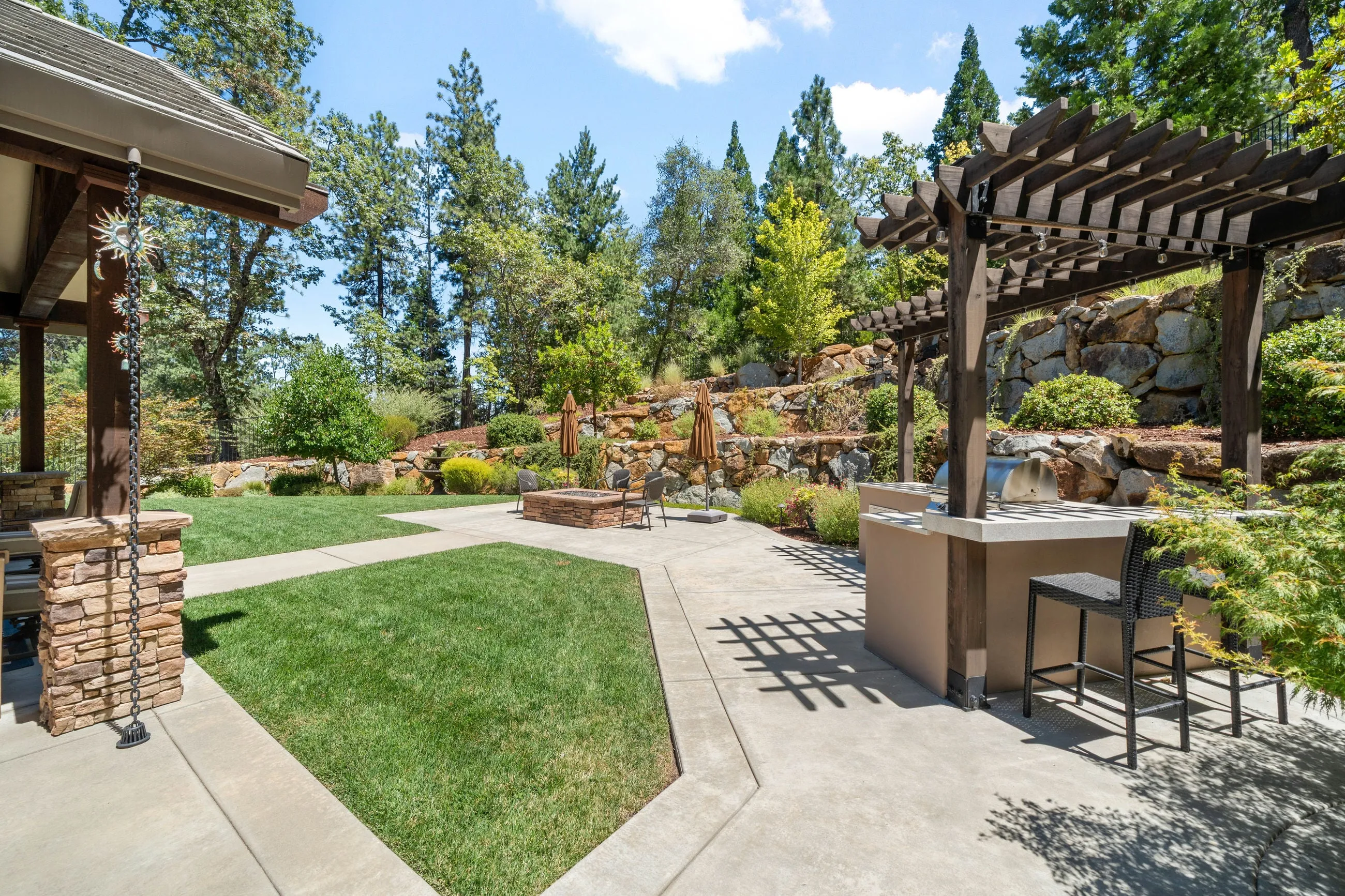 Lawn and patio with a pergola-covered outdoor kitchen, fire pit, and terraced boulder walls