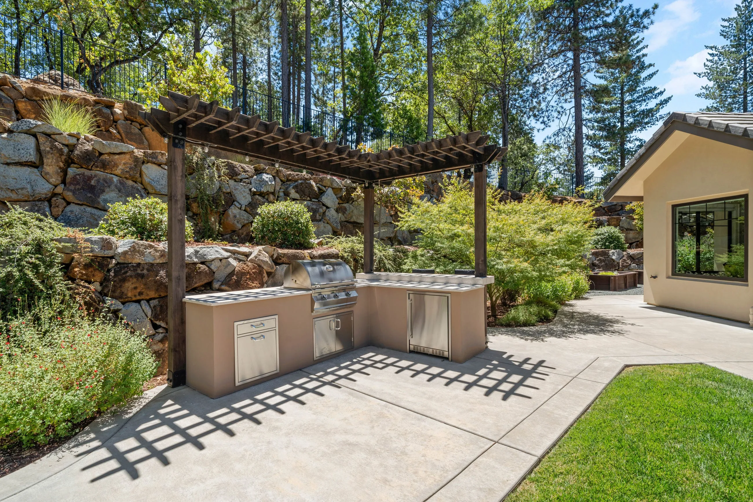 Outdoor kitchen under a timber pergola with built-in grill and fridge against the boulder wall