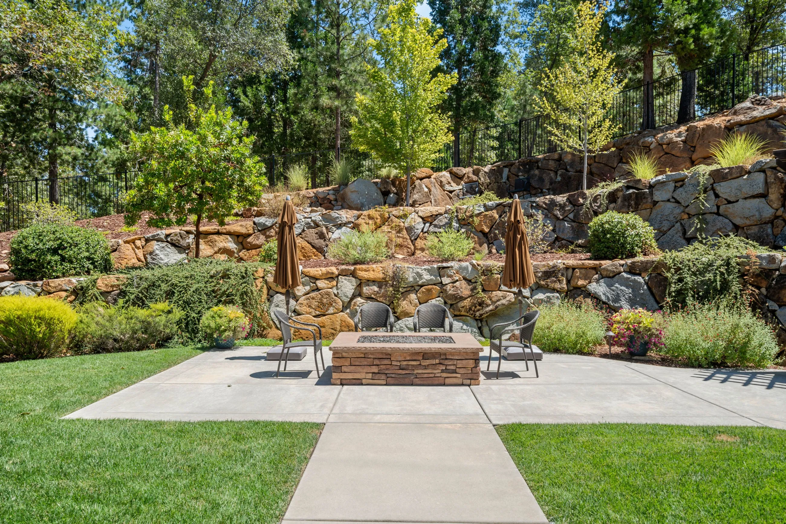 Square stone gas fire pit on the patio with chairs, umbrellas, and terraced rock gardens