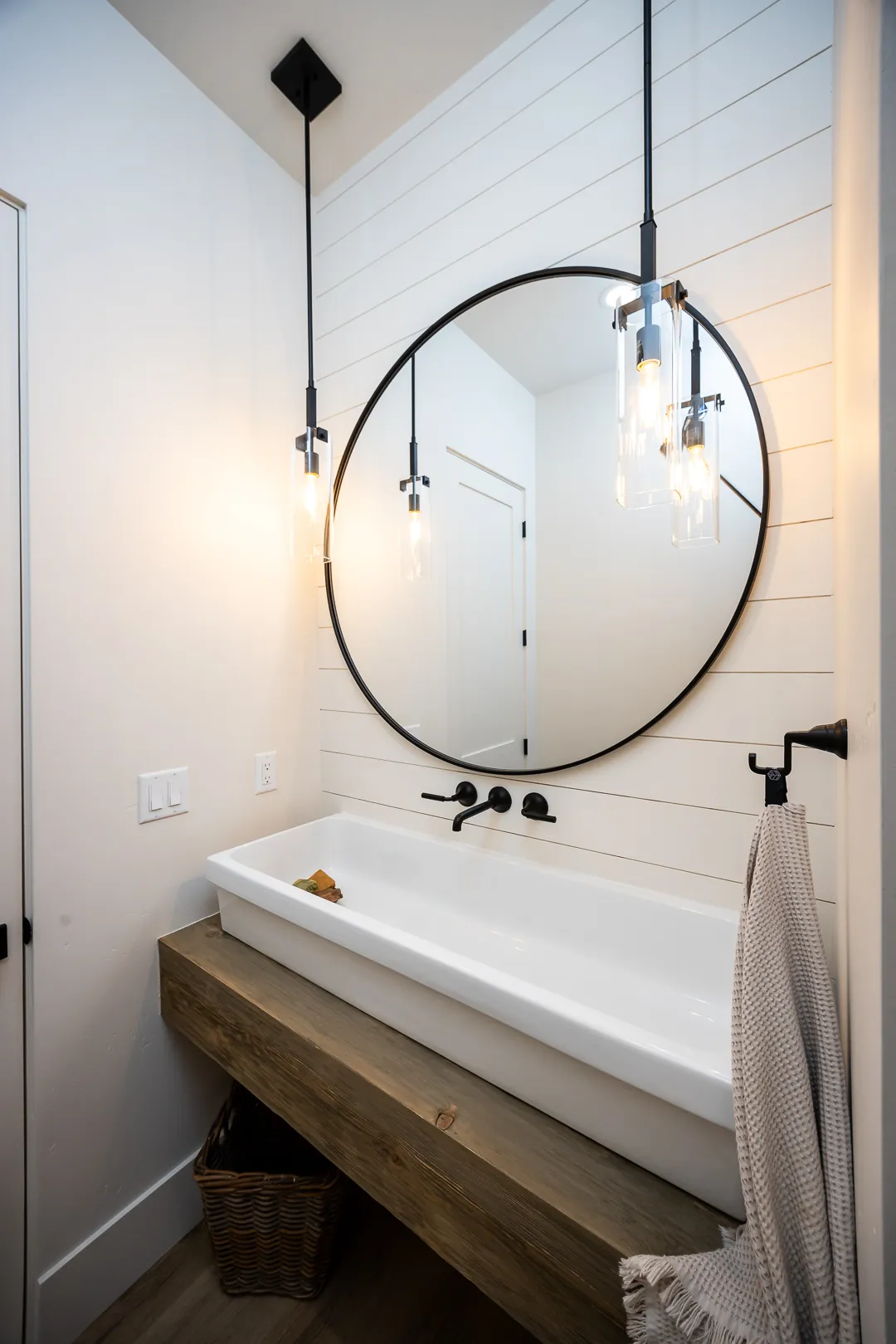Powder room with shiplap wall, round black-framed mirror, and trough sink on a floating wood vanity