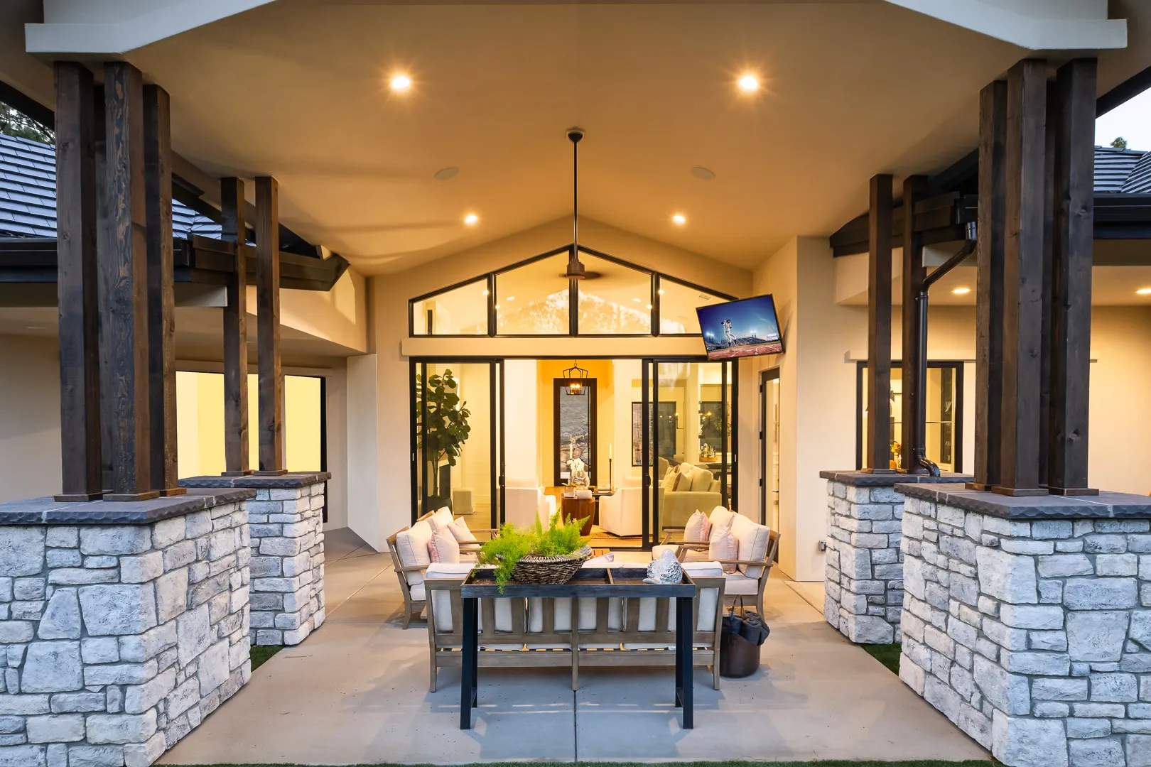 Evening view through the covered patio into the lit great room, with outdoor TV and dining