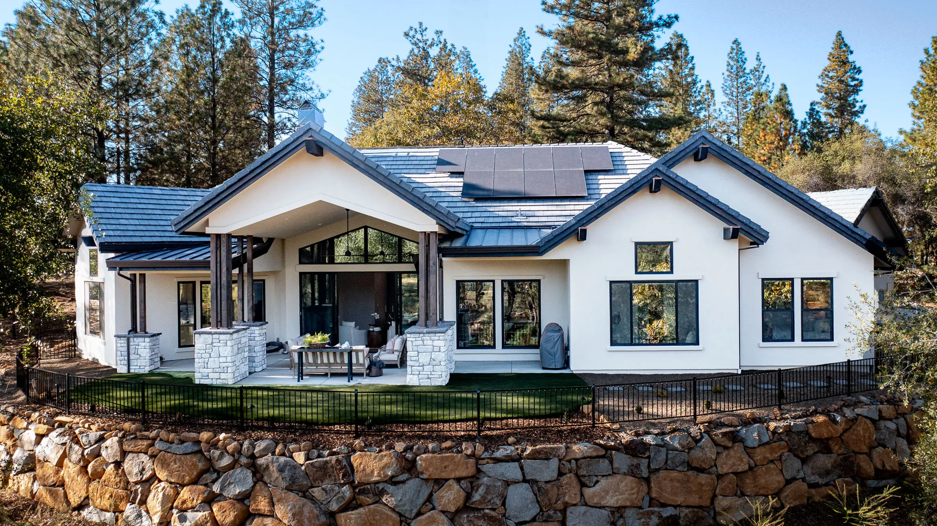 Rear exterior with solar panels, covered patio, fenced turf lawn, and natural rock retaining wall