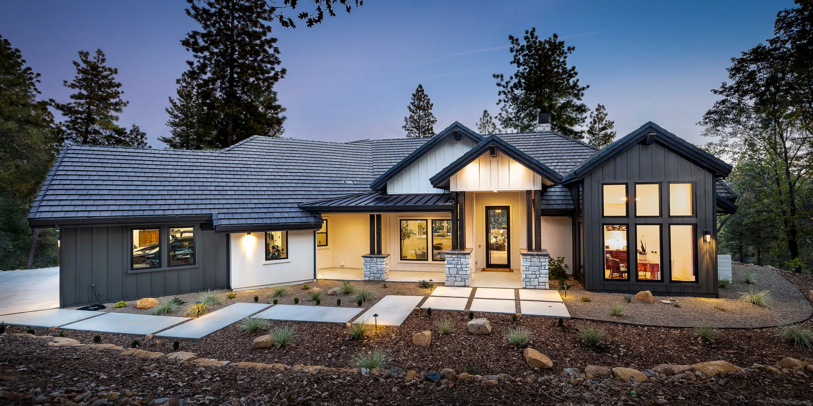 Front elevation of the Woodsy Elegance custom home at dusk, board-and-batten and stone among the pines