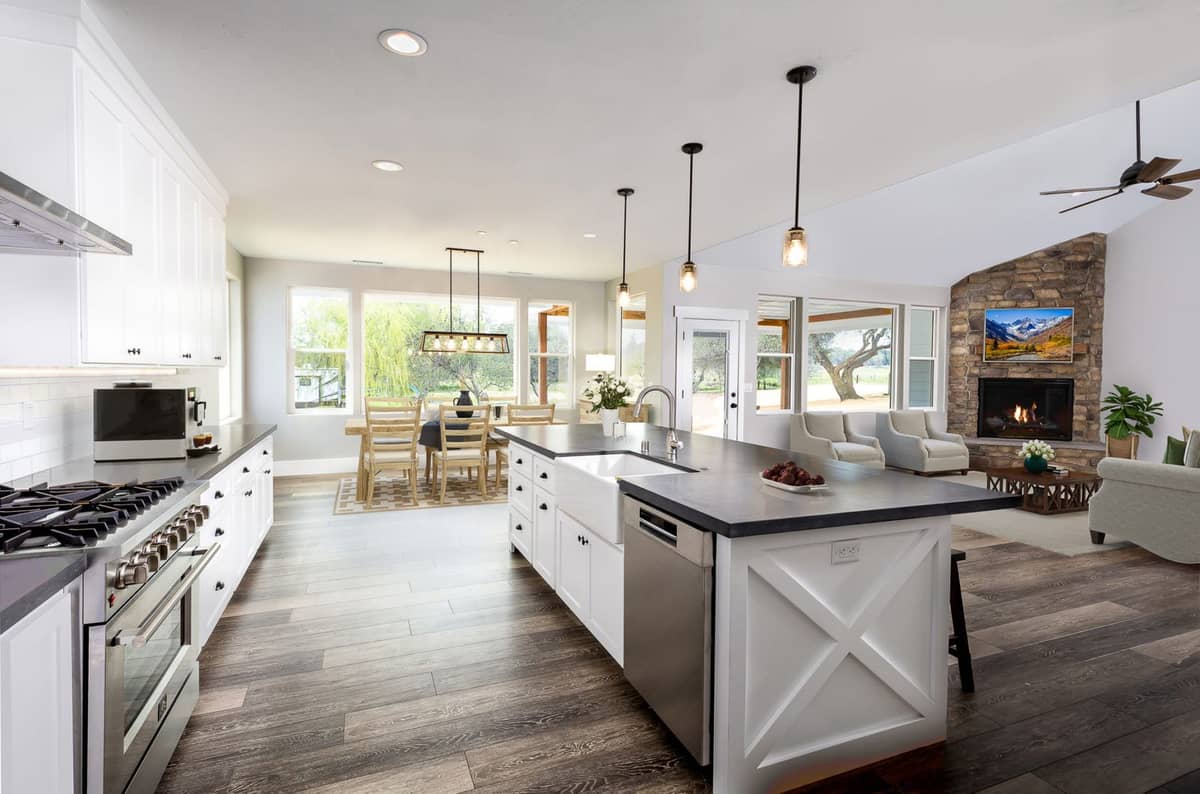 Open-concept farmhouse kitchen and great room with a white island, farmhouse sink, stone fireplace and dining area by the windows
