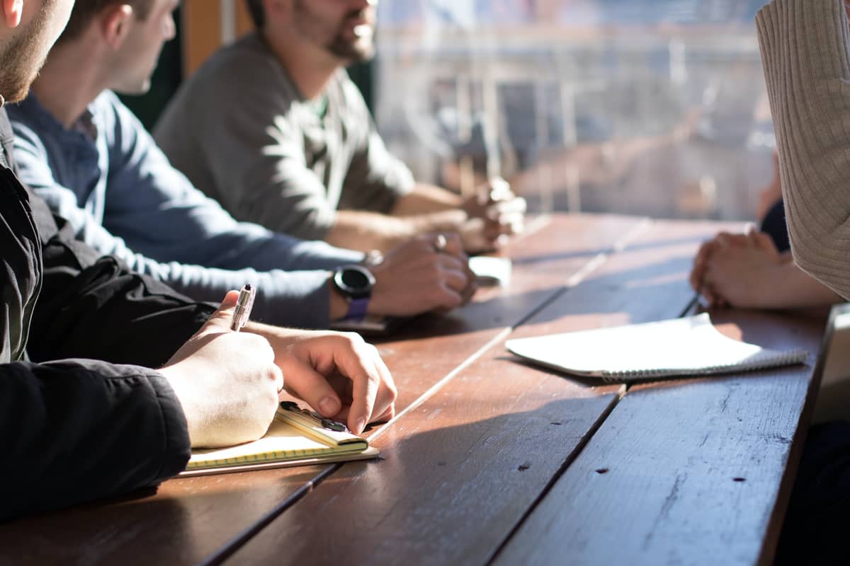 People taking notes during a planning meeting at a sunlit wooden table