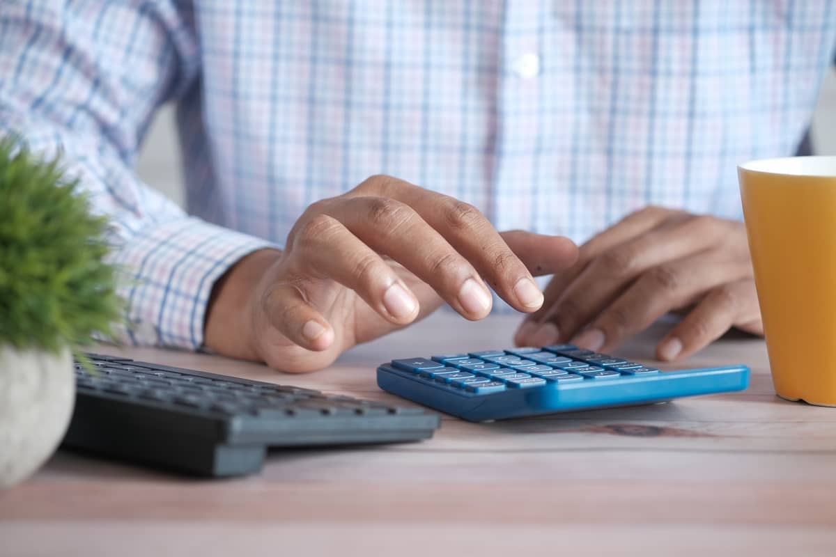 Close-up of hands tallying costs on a blue calculator beside a keyboard and coffee mug
