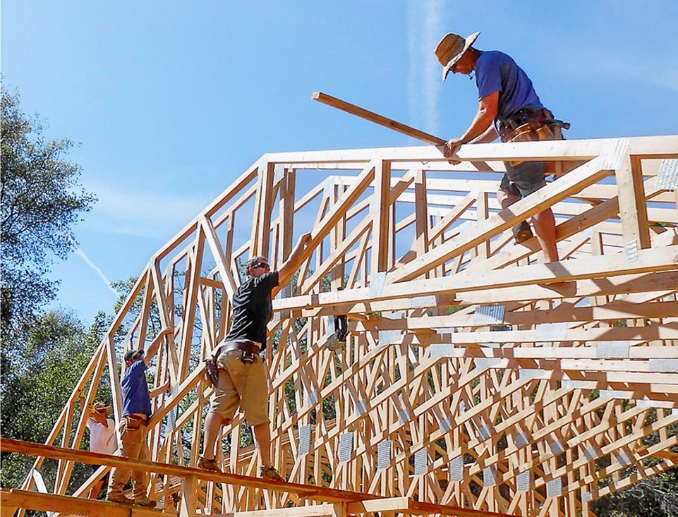 Framing crew setting wooden roof trusses on a new home under a clear blue sky