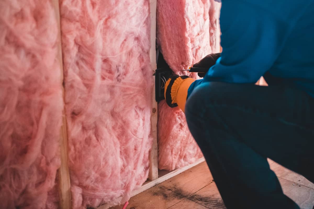 Worker wiring an outlet box in a wall filled with pink fiberglass insulation