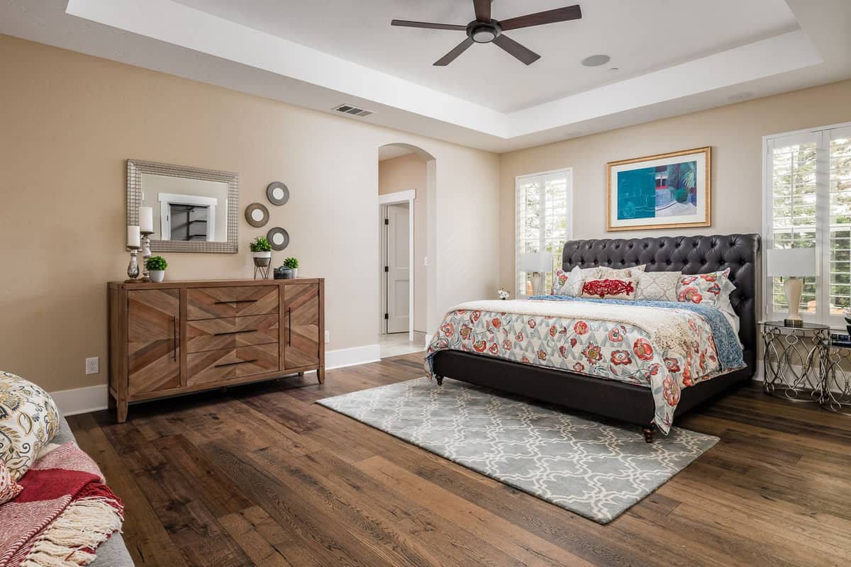 Spacious primary bedroom with tray ceiling, ceiling fan, tufted leather bed, and wide-plank floors
