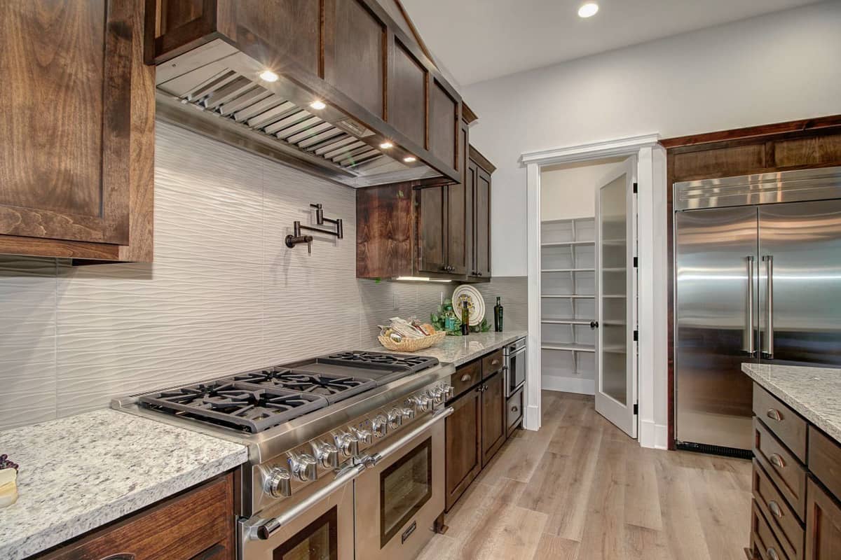 Kitchen with professional gas range, textured tile backsplash, and walk-in pantry beyond a glass door