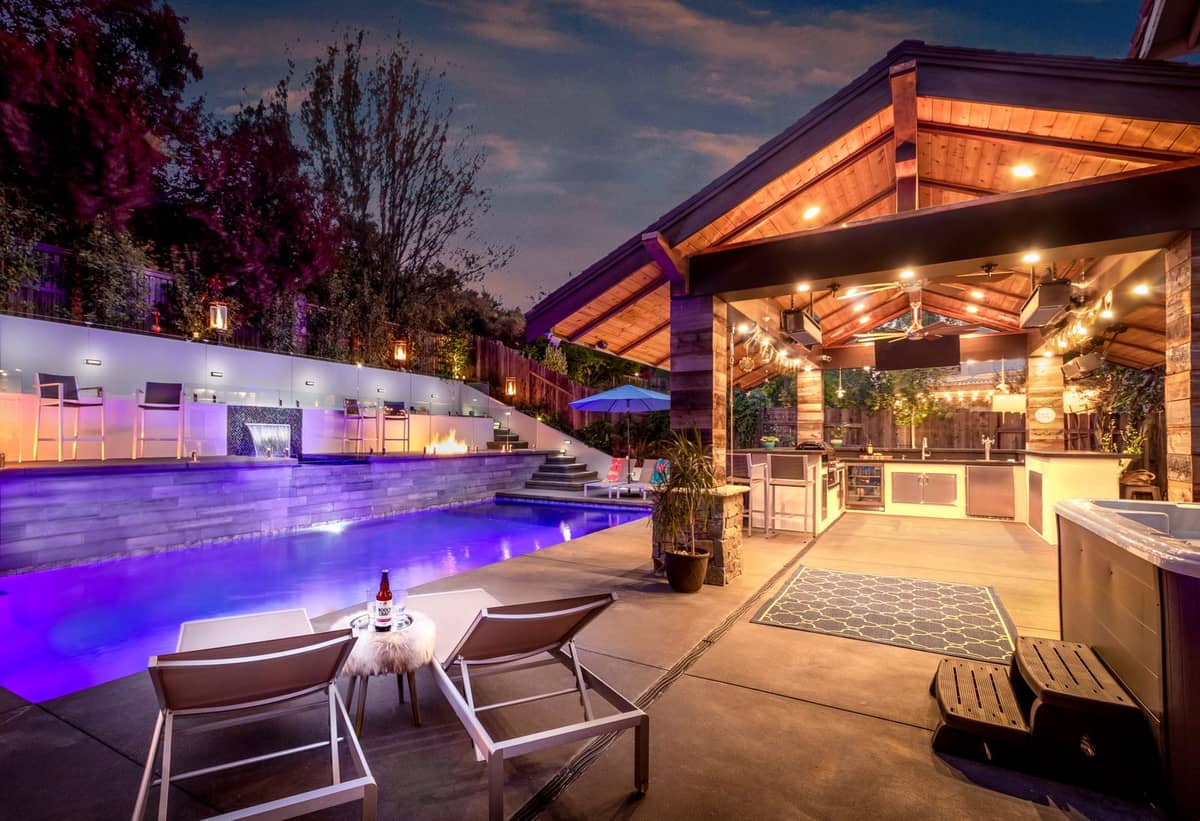 Backyard at dusk with lighted pool, lounge chairs, and covered outdoor kitchen pavilion